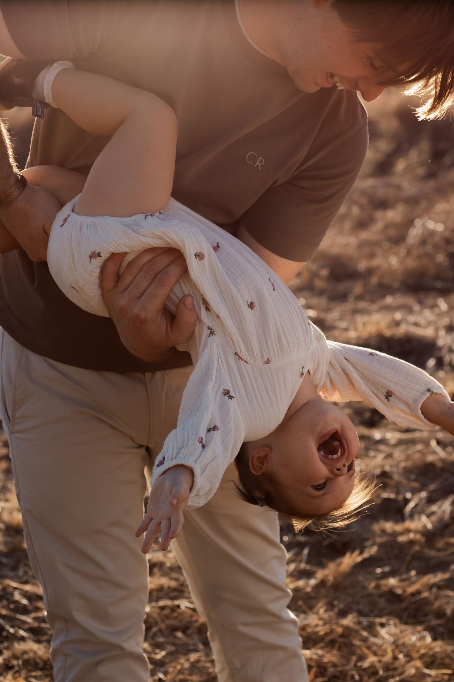 Parent playfully holding laughing baby upside down in a sunny outdoor field setting.