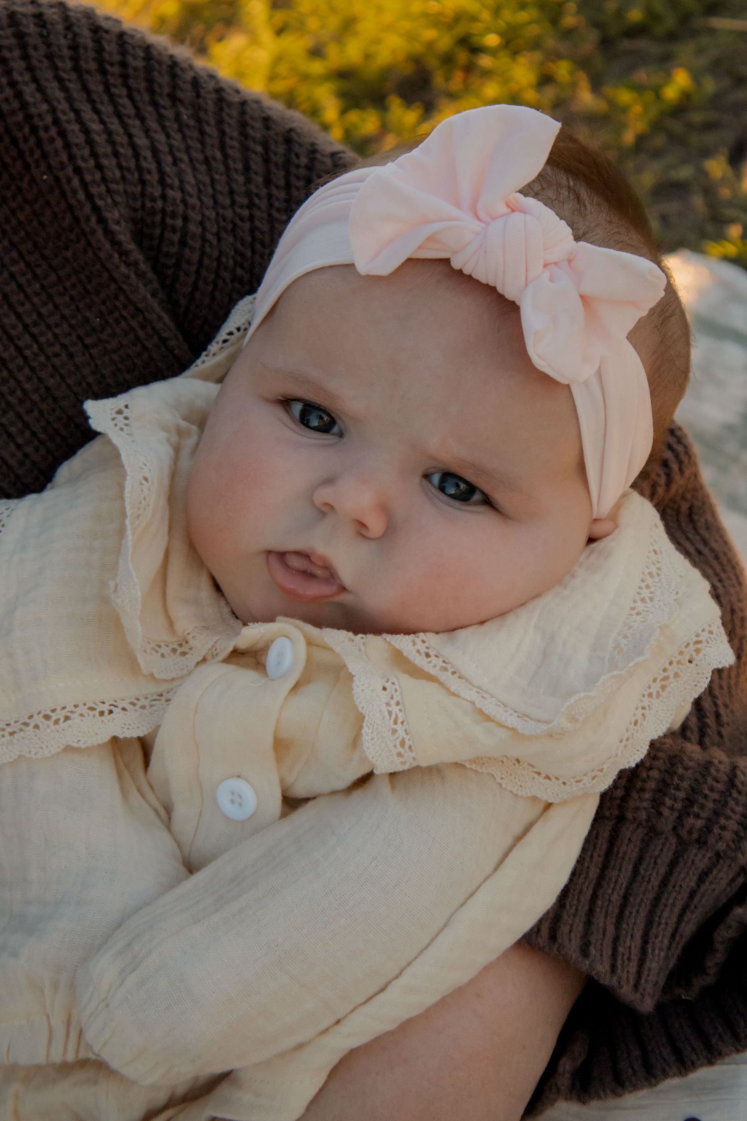 Adorable baby girl wearing a pink bow headband, wrapped in cream muslin cloth, being held outdoors in warm golden light.