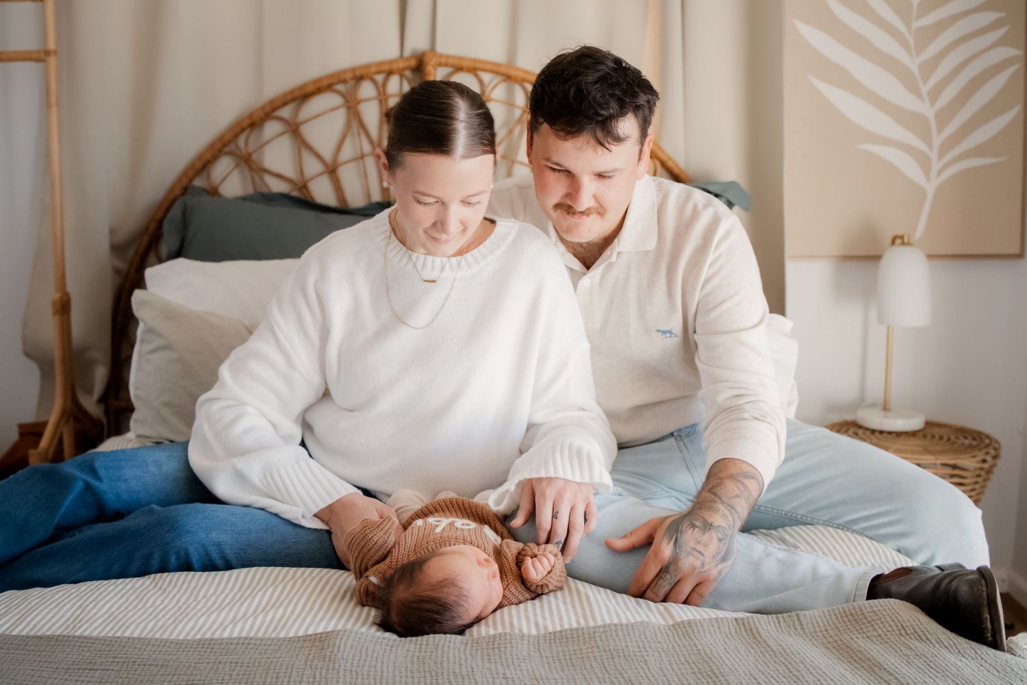 Happy young parents in white sweaters sitting on bed smiling at their newborn baby lying between them.