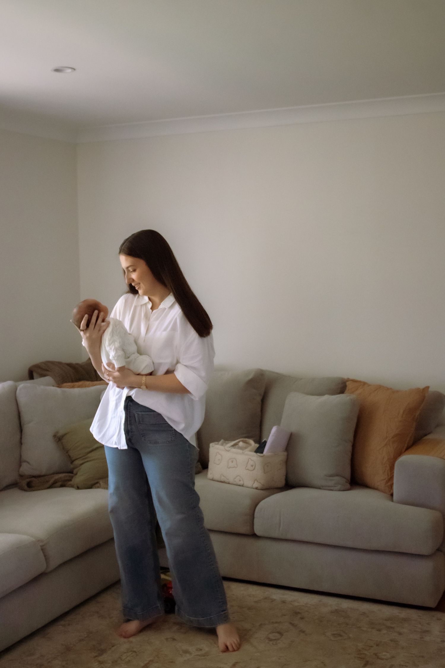 A young woman in a white shirt and jeans holds a newborn baby while standing in a cozy living room.
