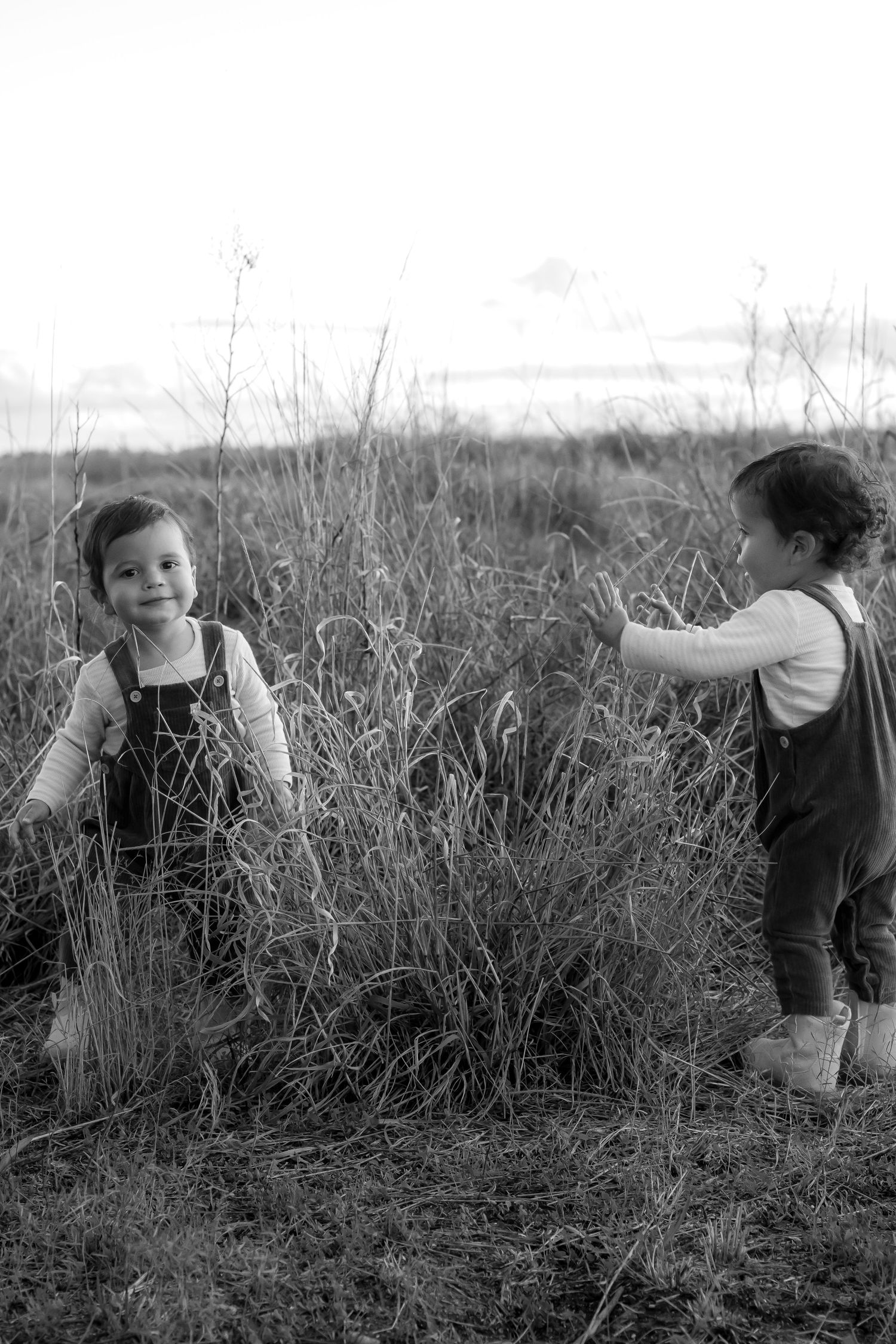 Two young children in overalls playing in tall grass fields, black and white photography.
