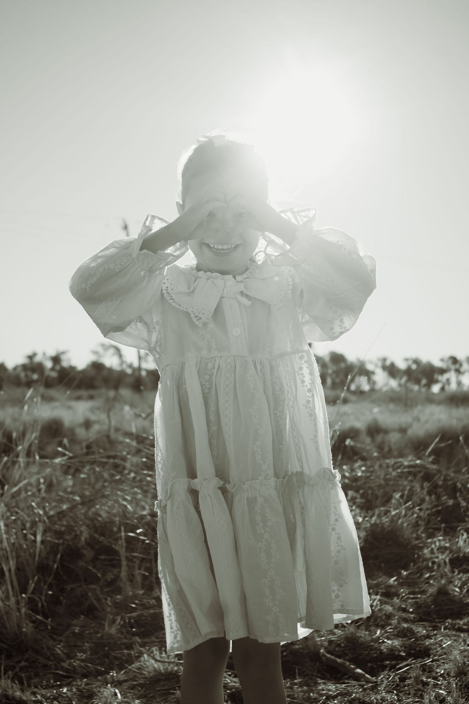 Girl in white lace dress standing in field, shielding eyes from bright sun, black and white photo.