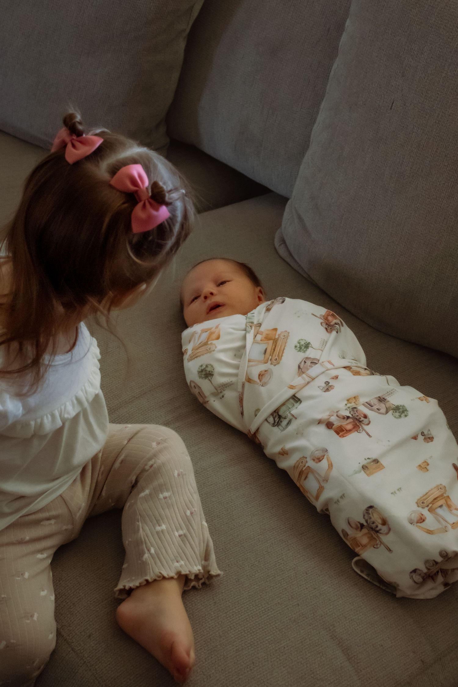 Young girl with pink bows looks at swaddled newborn baby lying on gray couch.