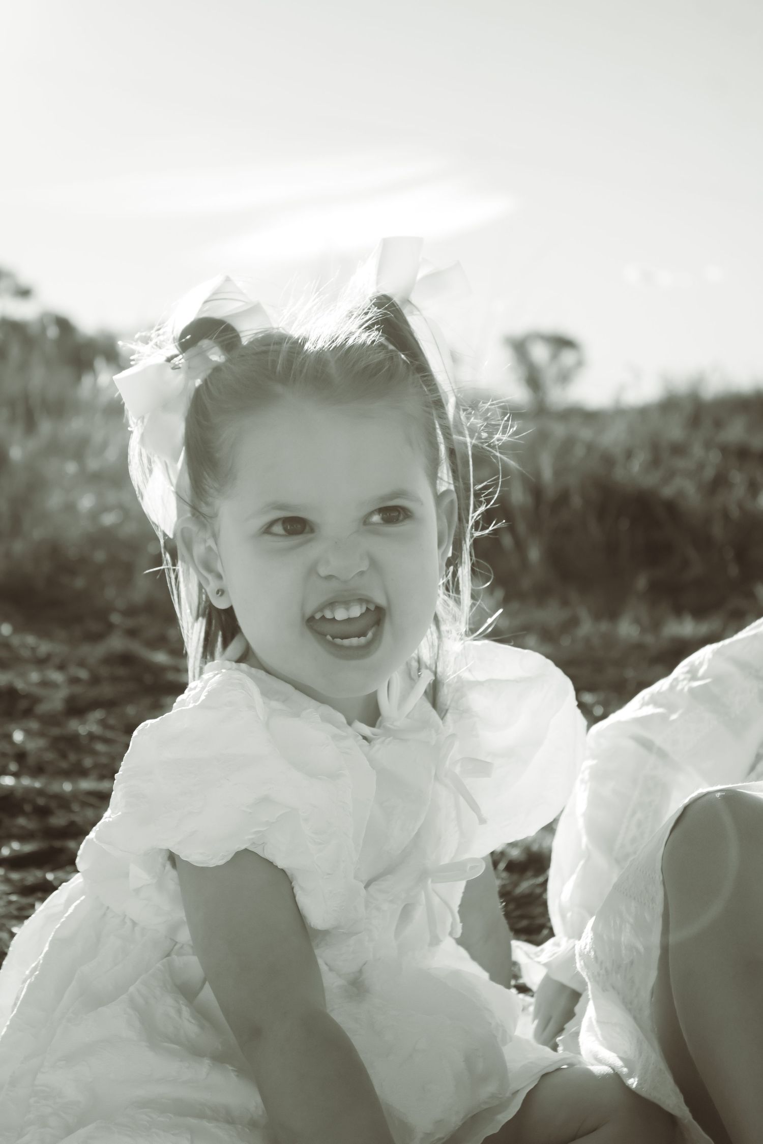 Black and white photo of a young girl in a white dress and bow, sitting outdoors with a playful expression.