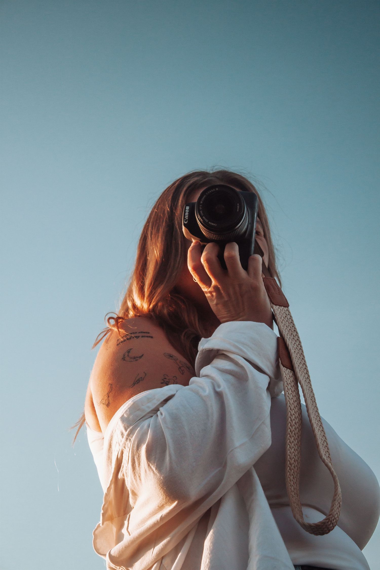 Woman with tattoos holding camera up to face against clear blue sky from below.