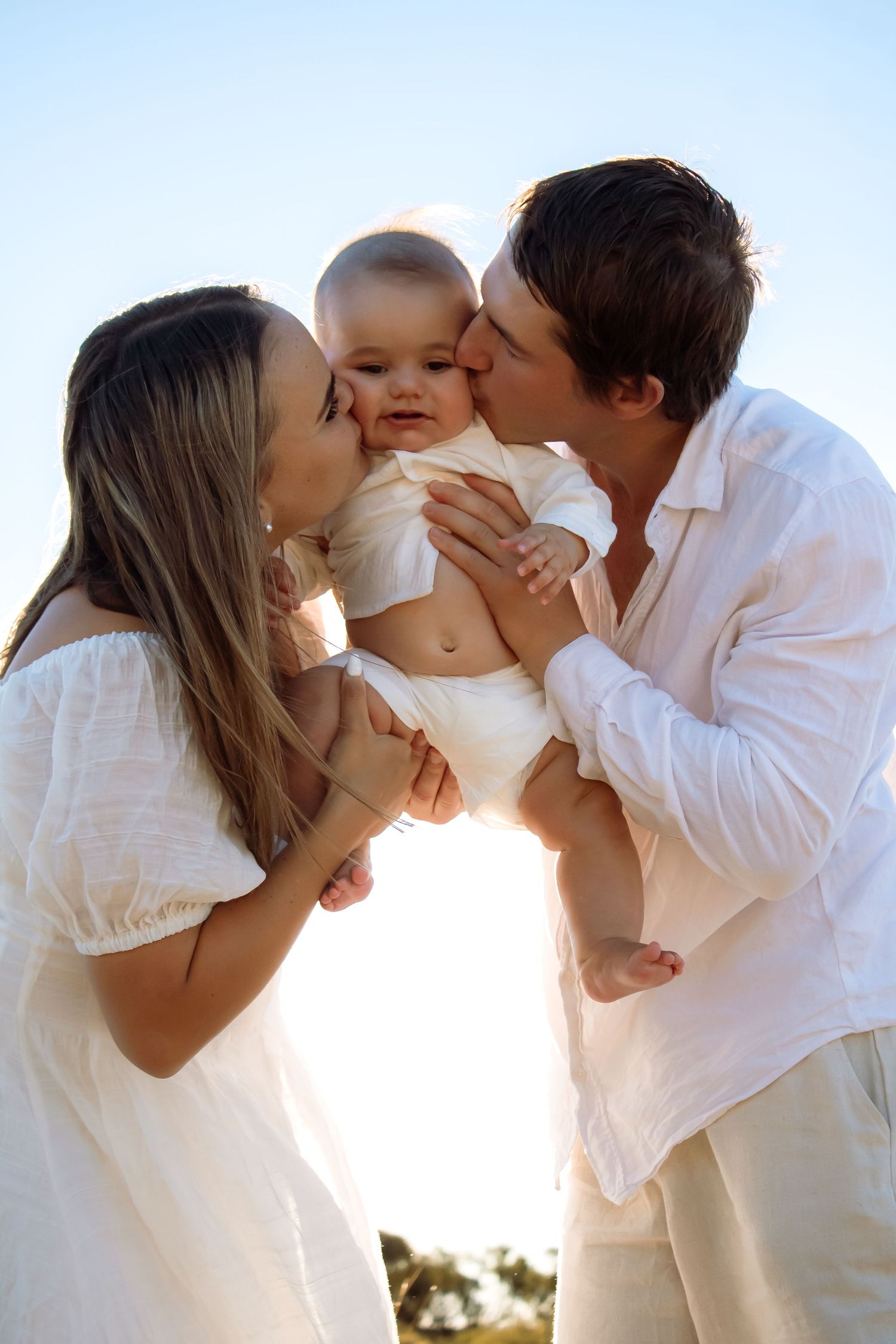 Parents in white outfits kissing their smiling baby outdoors against a clear blue sky.