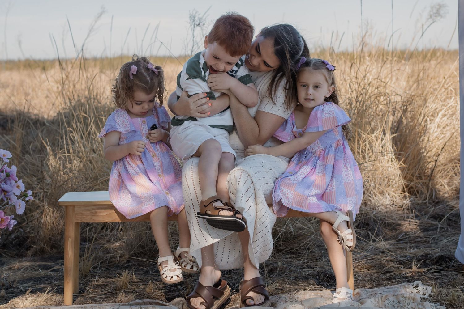 Family of five sitting on a bench in a golden field, with children in pink dresses and a boy in white.
