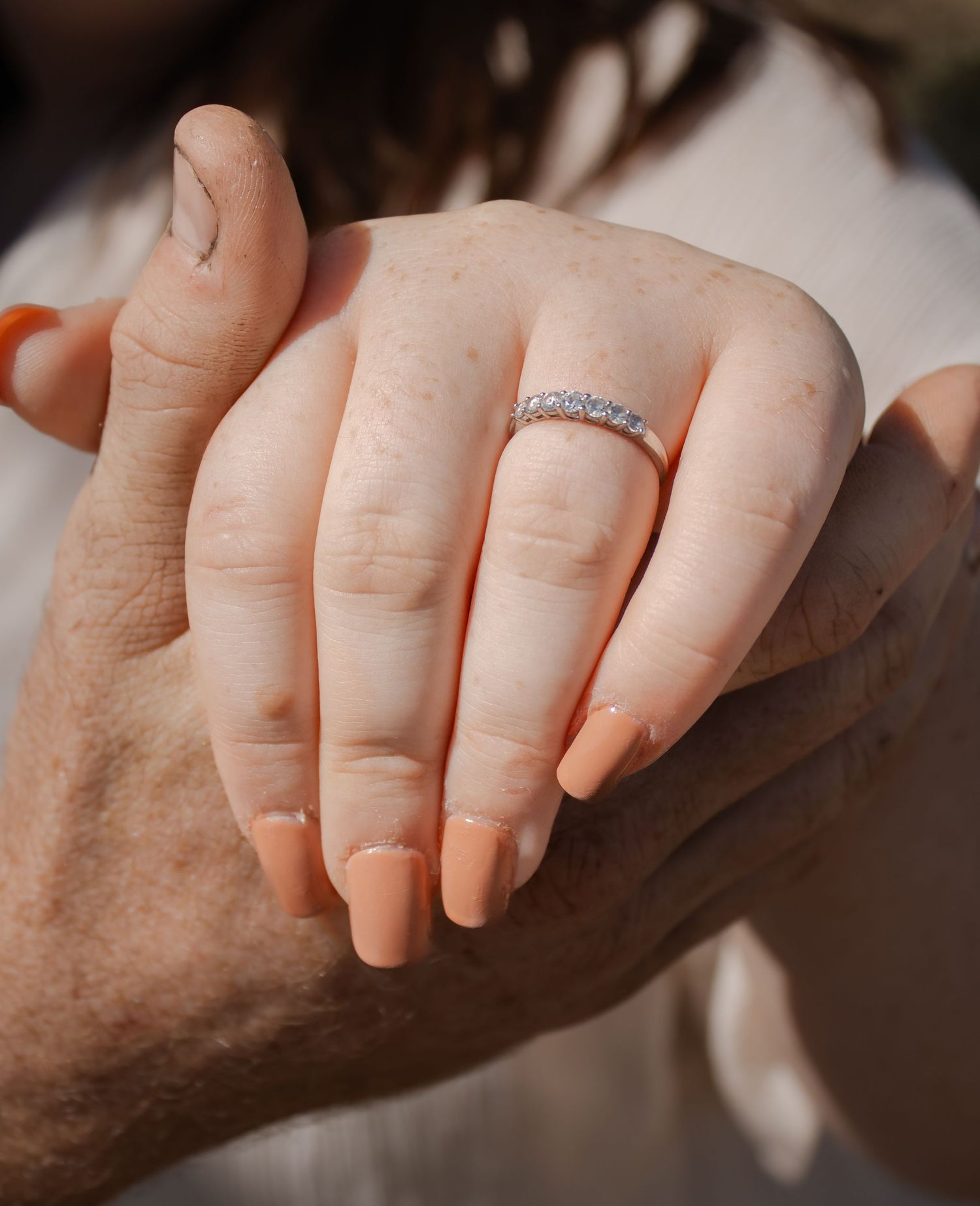 A young woman with peach nails wearing a diamond band ring holds hands with an older person in warm sunlight.