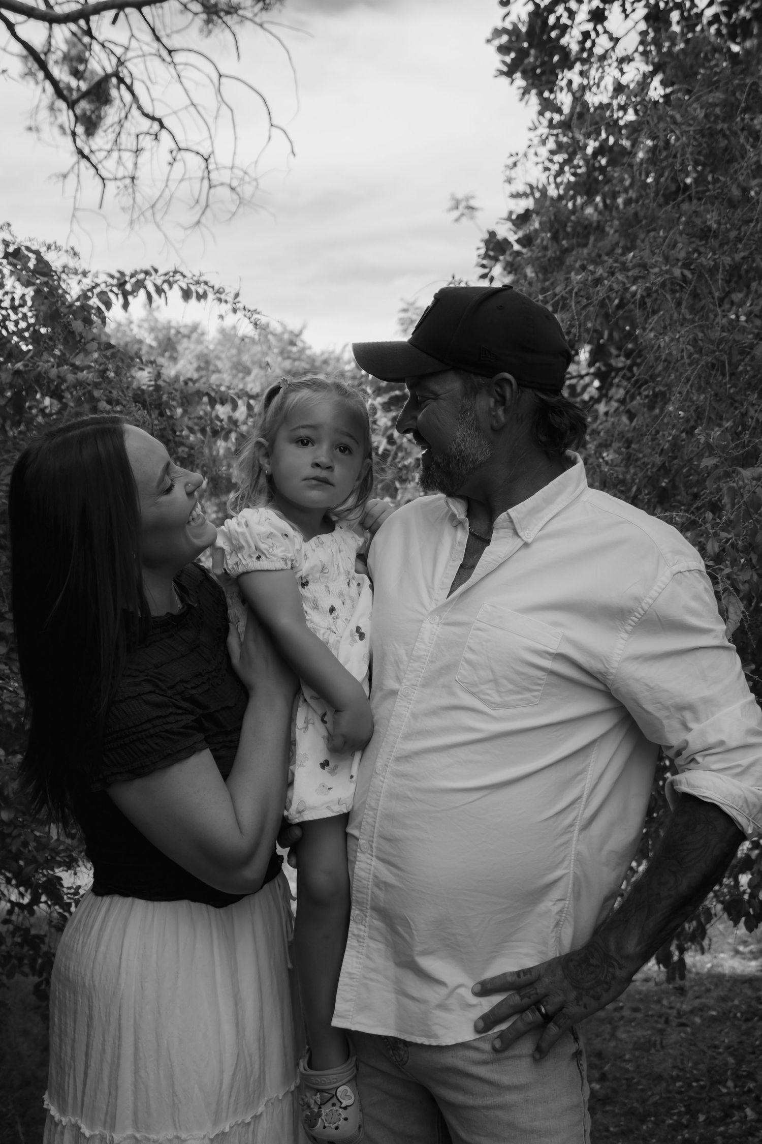 Black and white family portrait of a woman, young girl, and older man in a cap outdoors among trees.