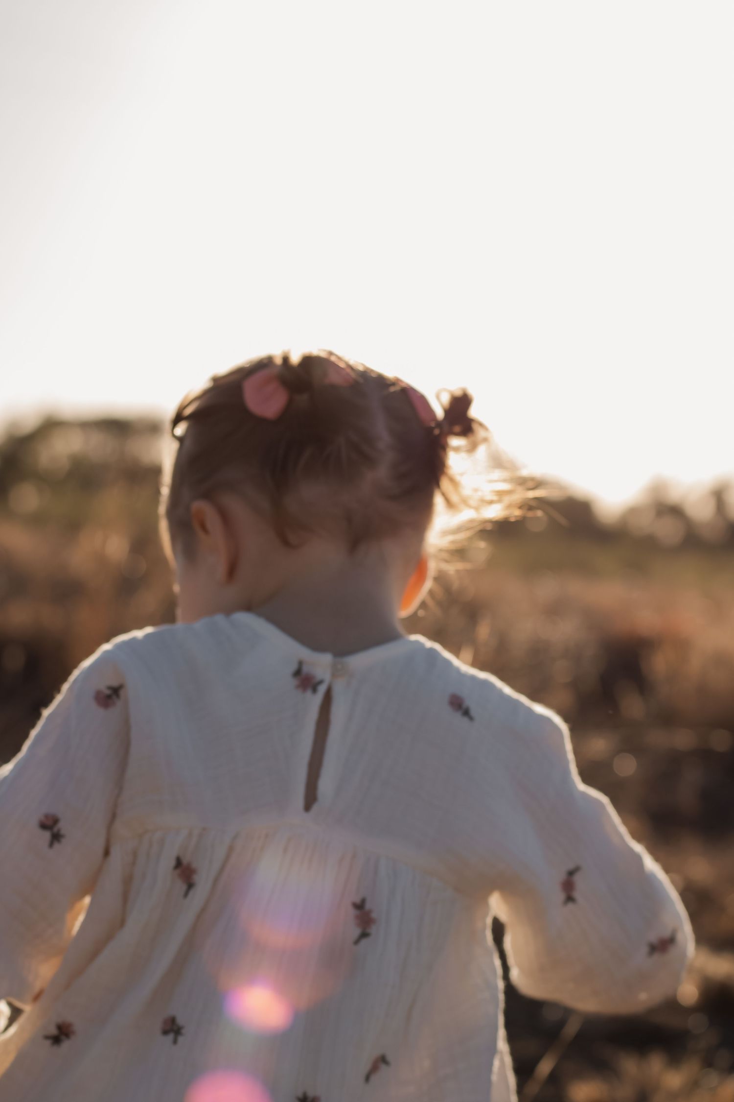 A toddler girl in a white embroidered dress with pigtails runs through a sunlit field at golden hour.