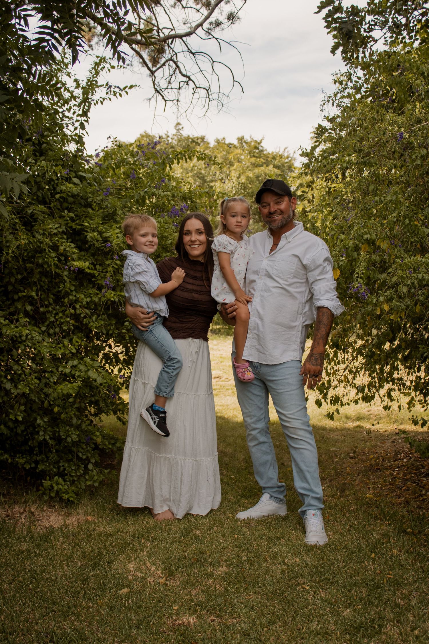 Happy family of four posing outdoors under lush green trees, parents holding young children in natural light.