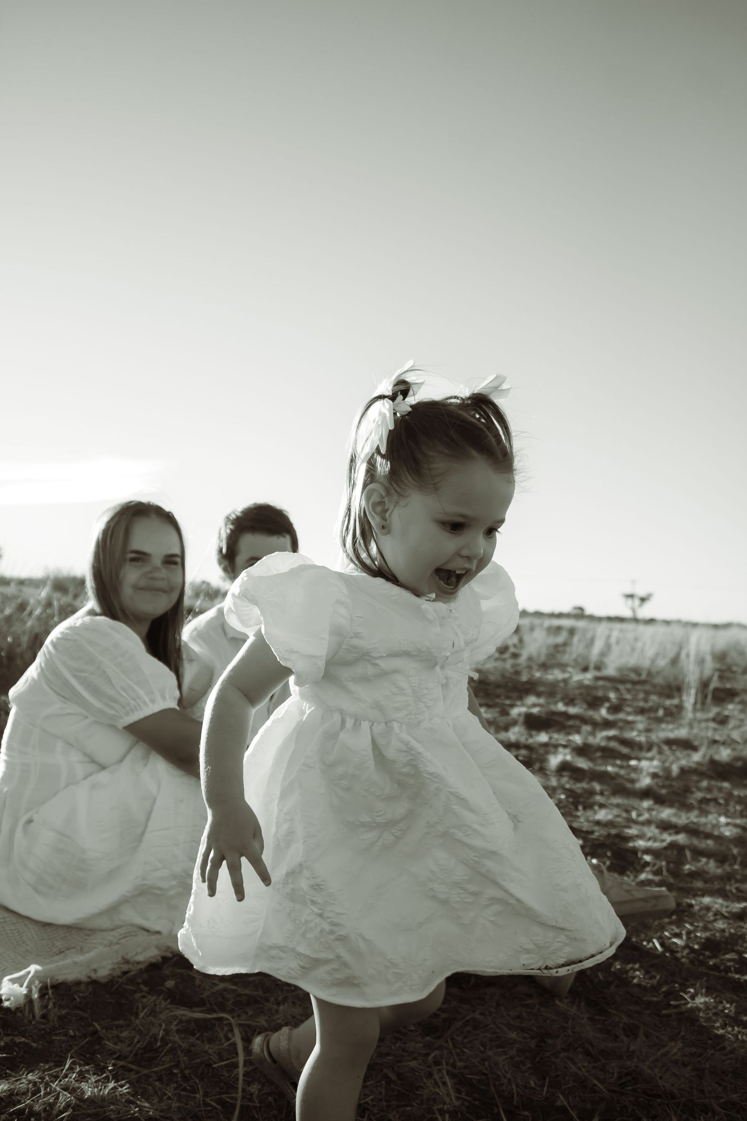 Young girl in white dress runs joyfully outdoors in black and white photo with siblings in background.