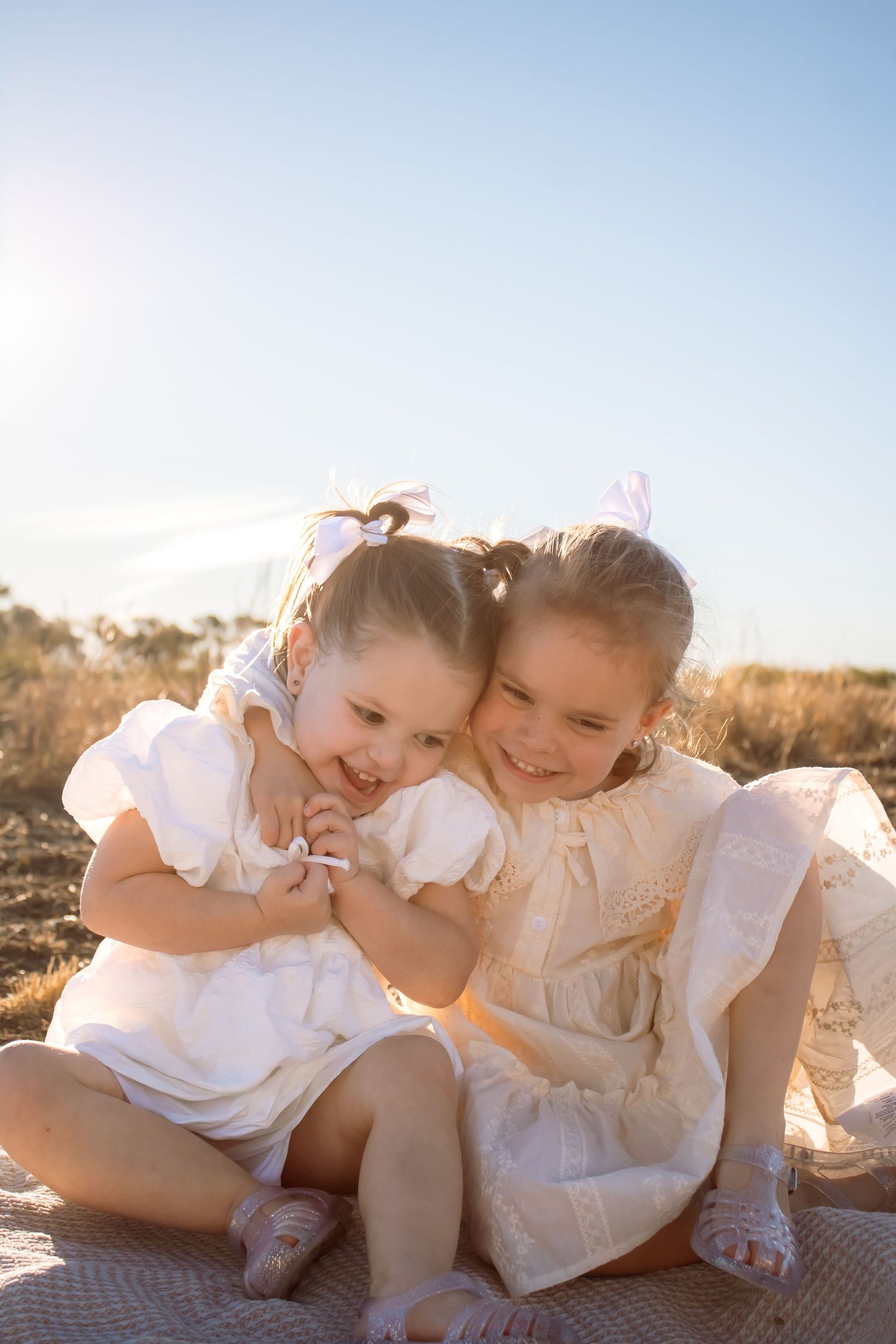Two young girls in white dresses with pink bows sit outdoors, hugging and smiling in golden sunlight.