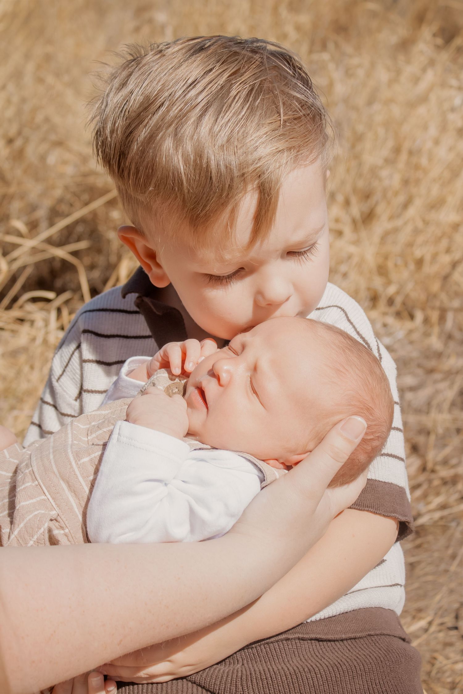 Young boy kissing sleeping newborn baby sibling held in adult hands outdoors in golden field.