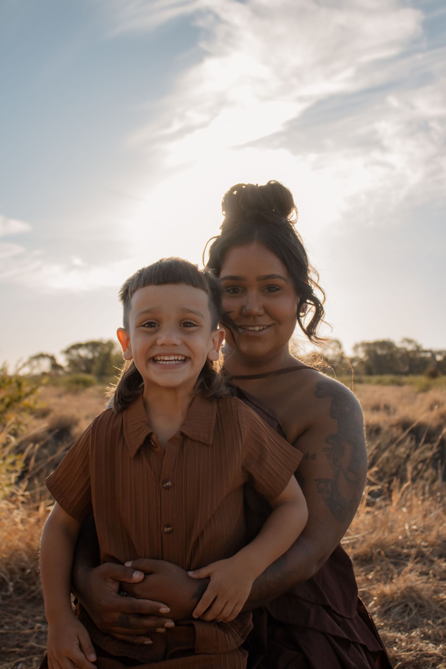 A woman and young boy smile together in a sunlit field, wearing matching brown outfits in an outdoor portrait.