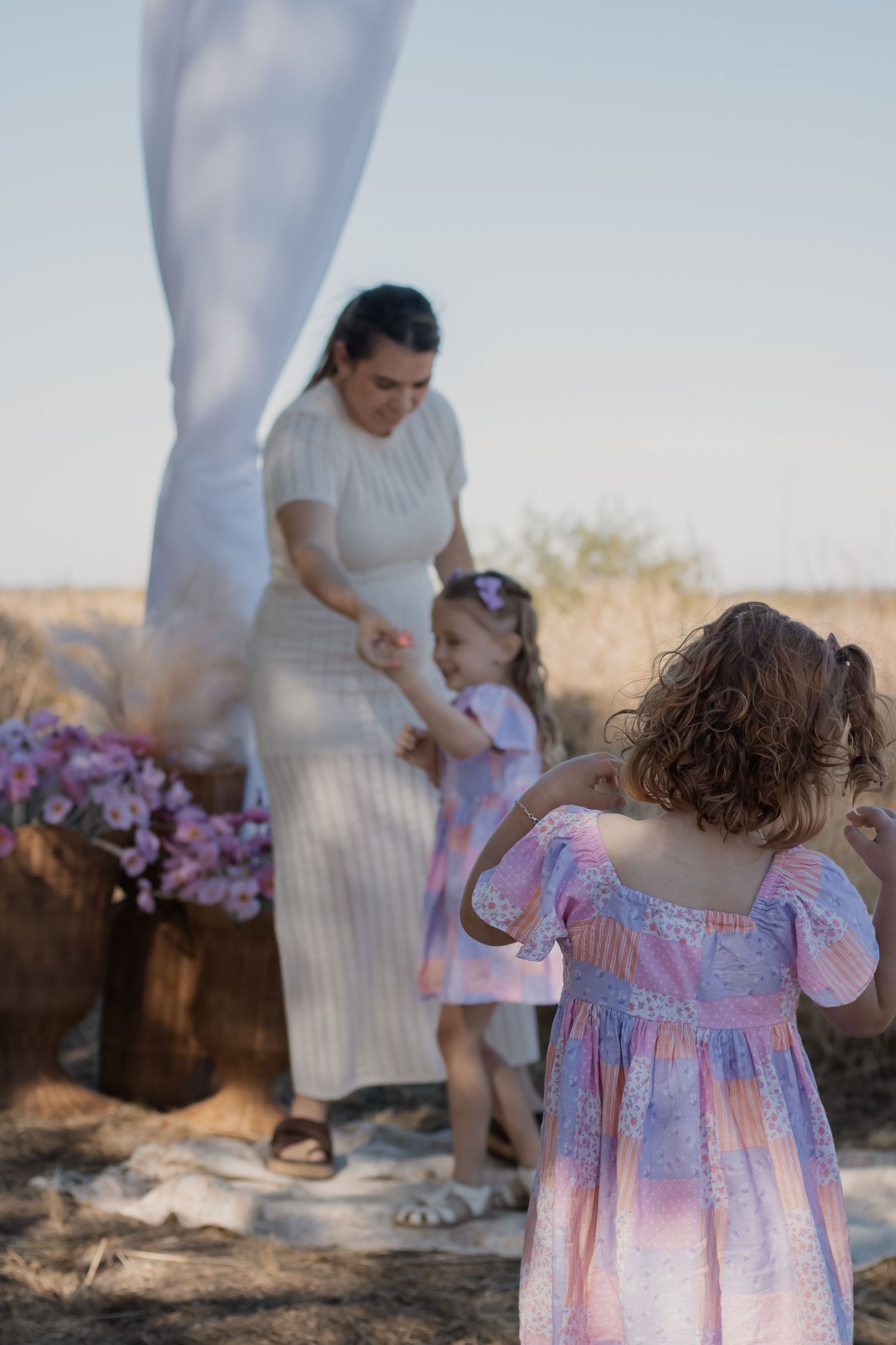 A woman in white dress holds hands with a little girl at an outdoor floral setup while another curly-haired girl watches.