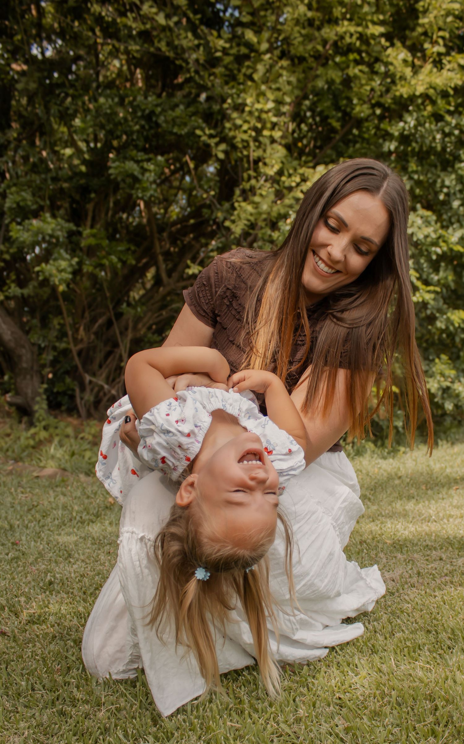 A smiling mother playfully holds her laughing blonde toddler upside down on green grass with trees in the background.