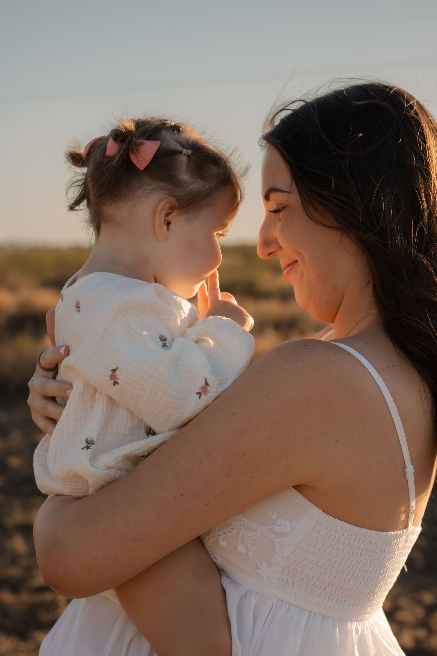 A smiling mother in a white dress holds her toddler daughter outdoors at golden hour sunset.