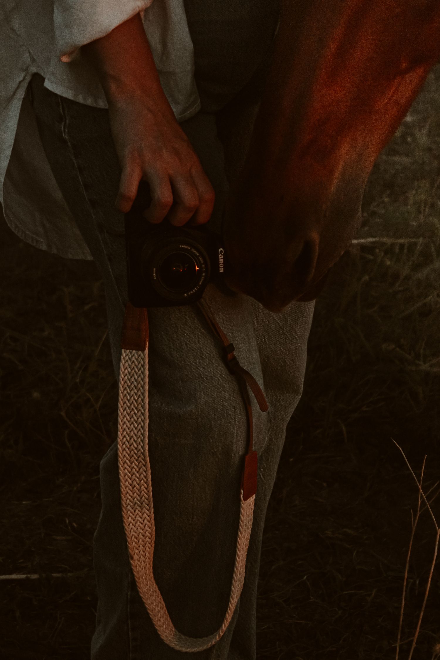 Person holding a Canon camera with a braided strap, wearing jeans, photographed in low light outdoors.