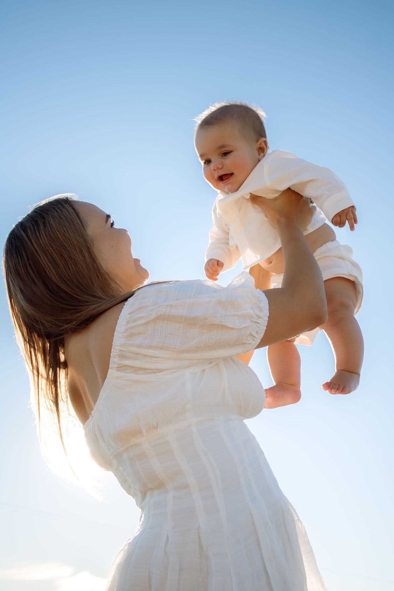 A joyful mother in a white dress lifts her smiling baby against a bright blue sky.