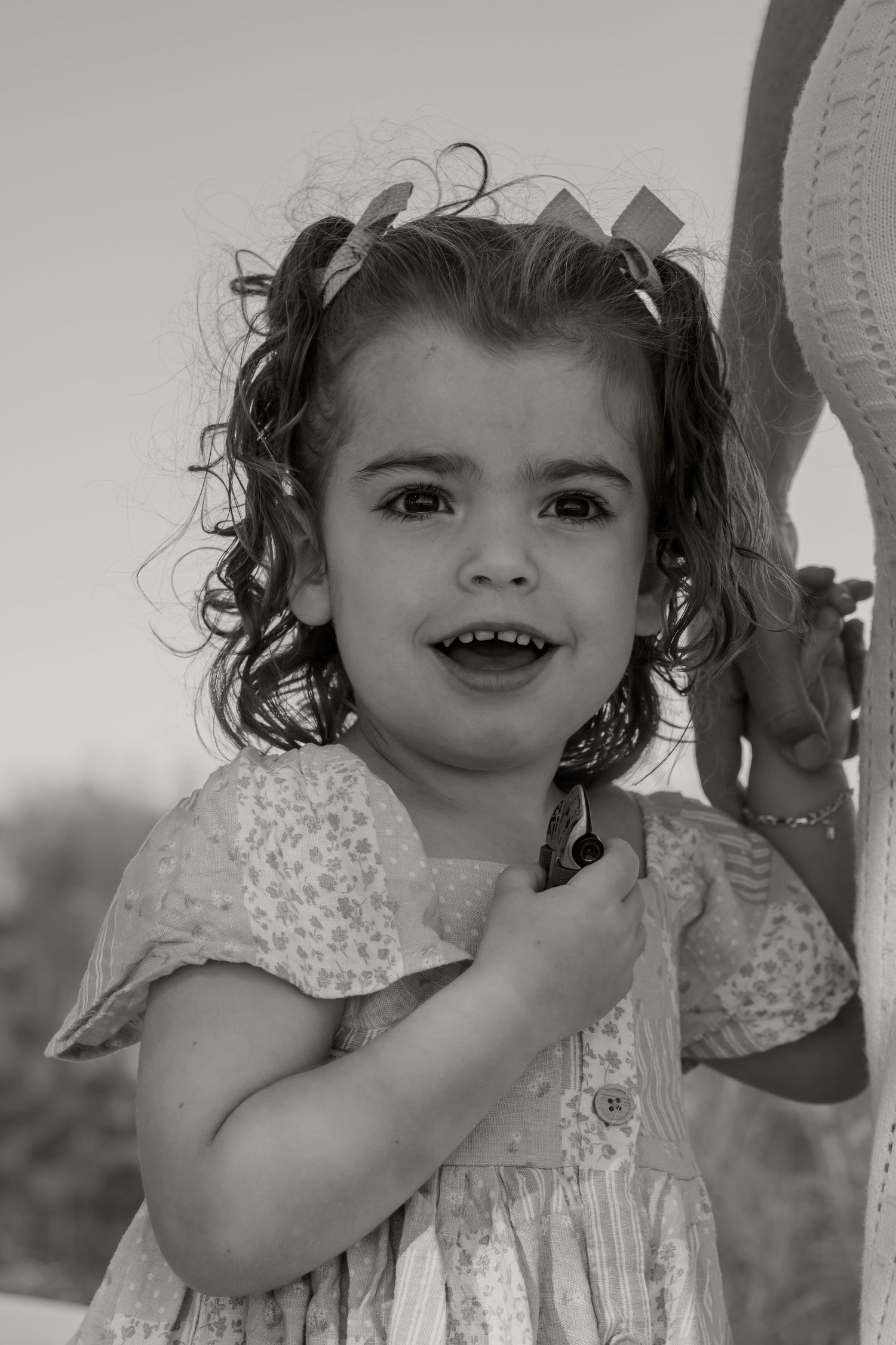 Black and white portrait of a smiling toddler girl with curly pigtails wearing a floral dress.