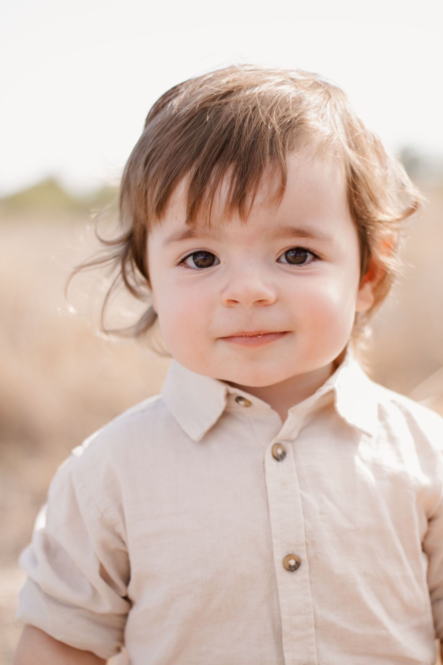 Adorable toddler with brown eyes wearing a beige linen shirt, smiling outdoors in a sun-drenched field.