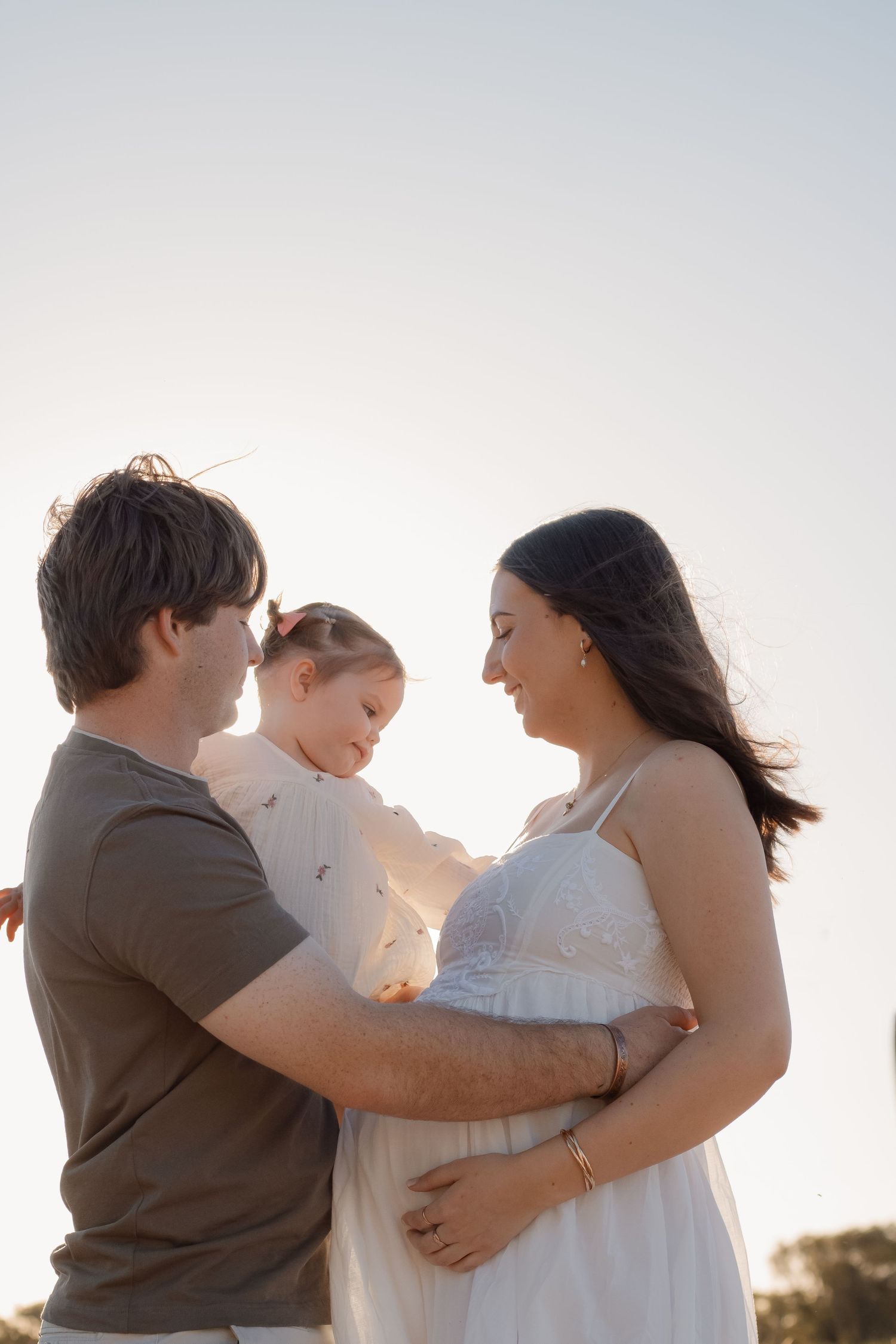 A couple holding their young child outdoors at golden hour, with the pregnant woman in a white dress.