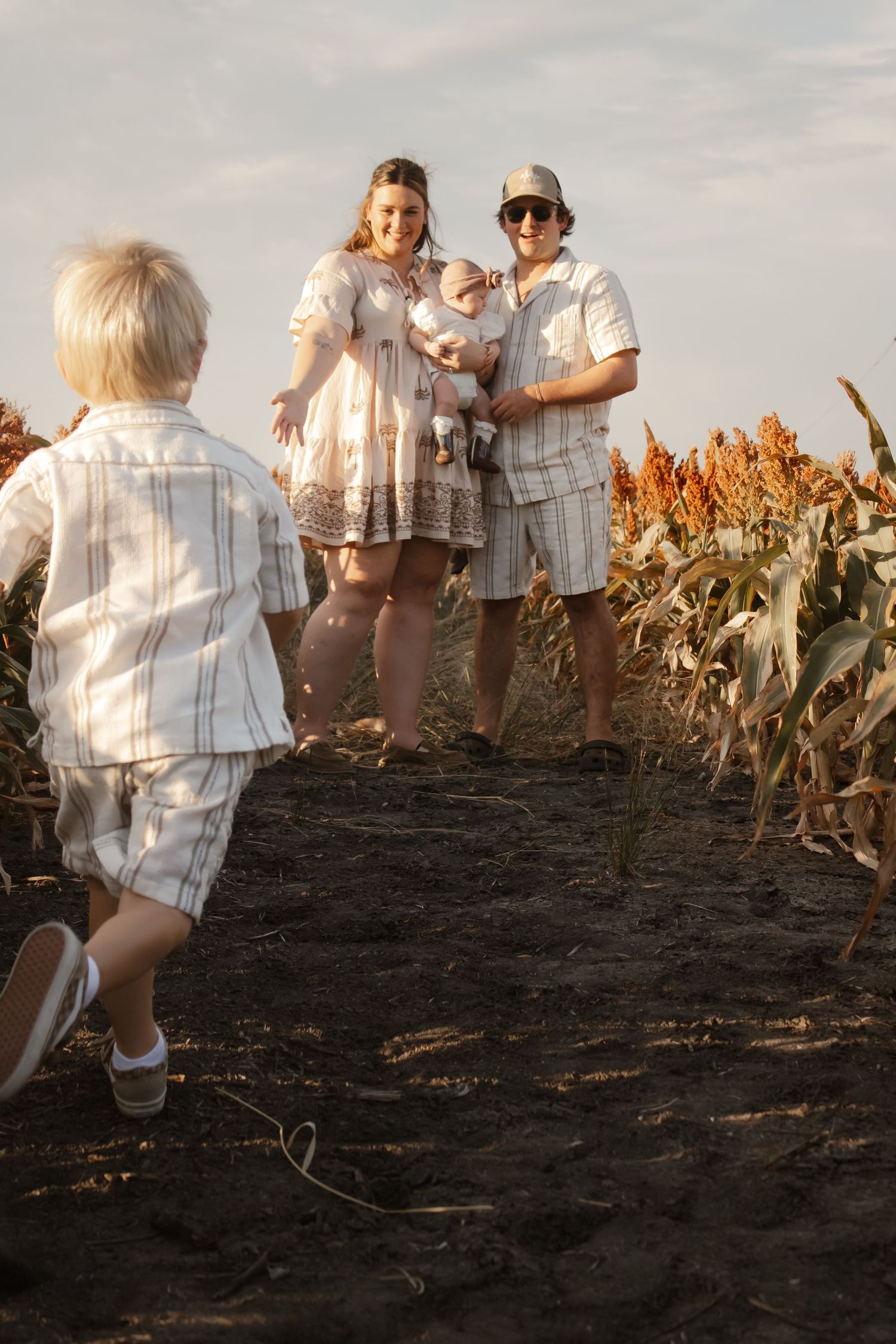 Family in a cornfield, parents holding baby while young child runs toward them on a sunny day.