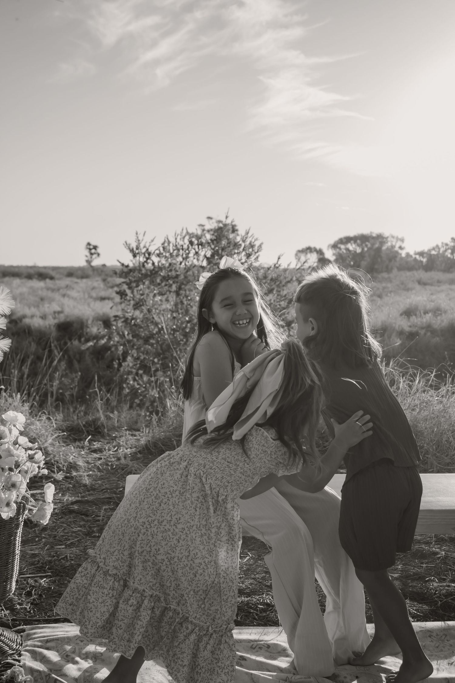Black and white photo of a woman in a floral dress hugging two young girls outdoors in a sunlit field.