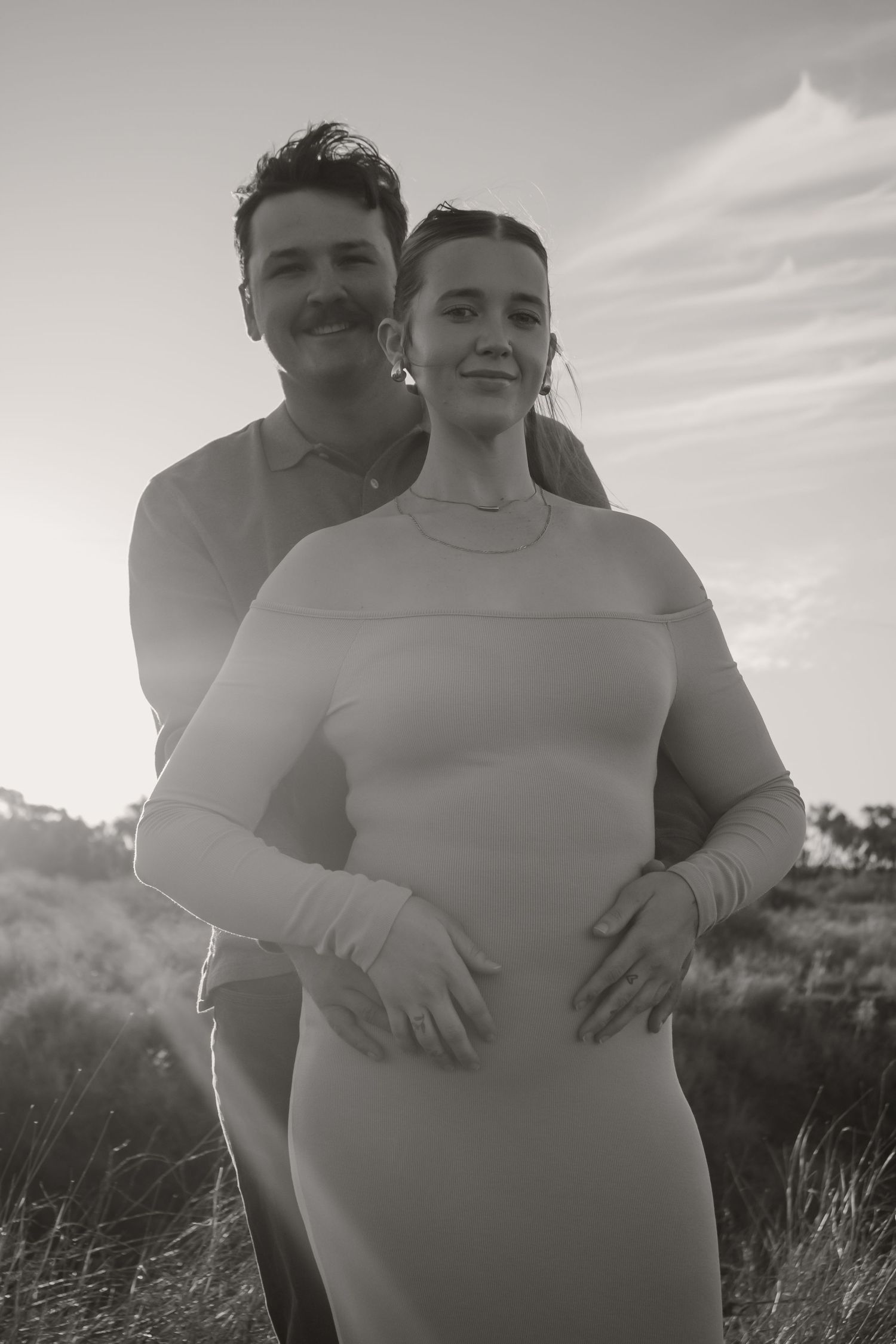 Black and white photo of a couple embracing outdoors, pregnant woman in fitted dress with partner holding her belly.
