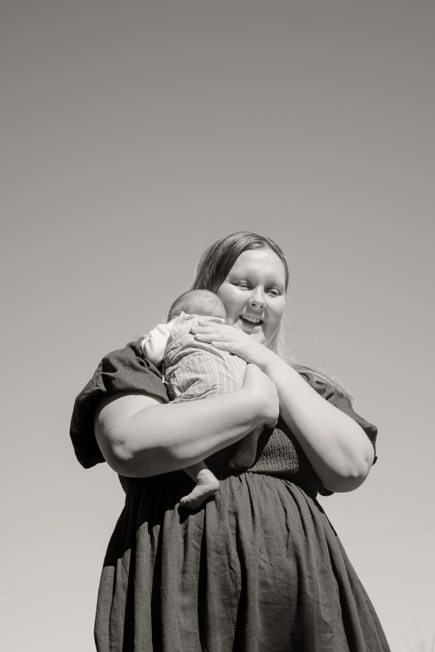 Black and white photo of a woman holding a newborn baby outdoors against a clear sky.