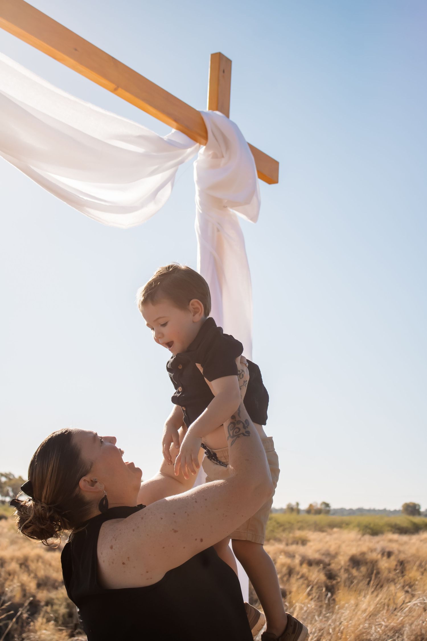 A mother joyfully lifts her toddler near a wooden cross with white fabric outdoors under a clear blue sky.