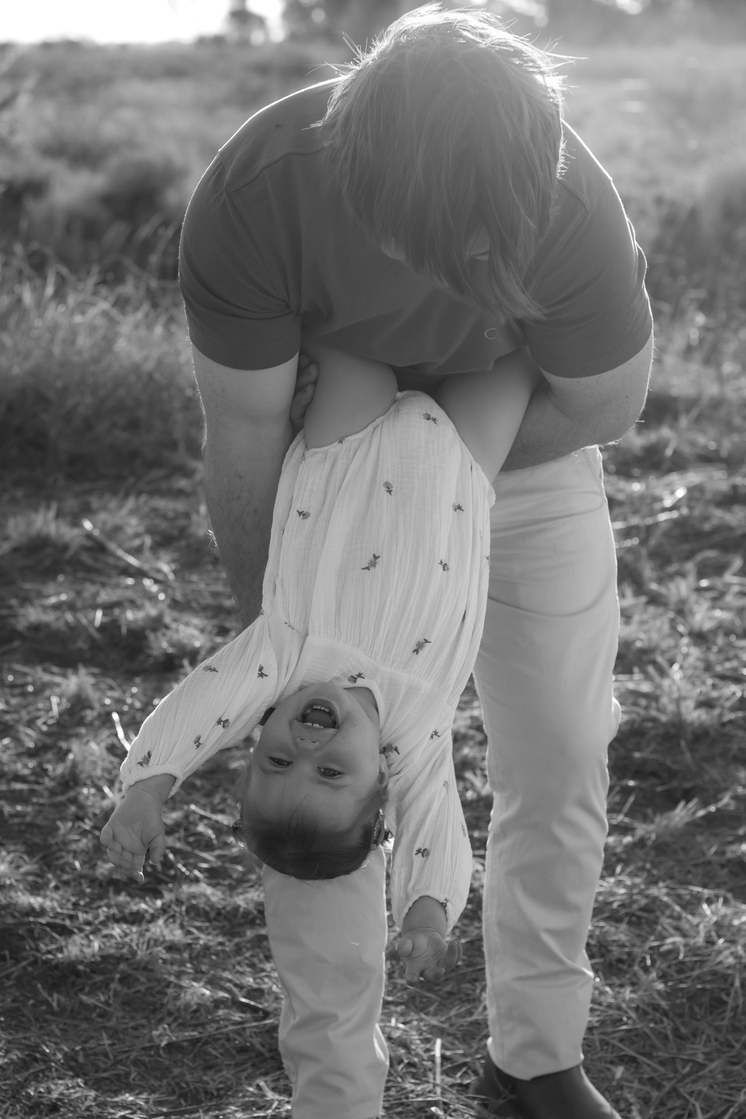 Black and white photo of a parent playfully holding a laughing child upside down in a grassy field.