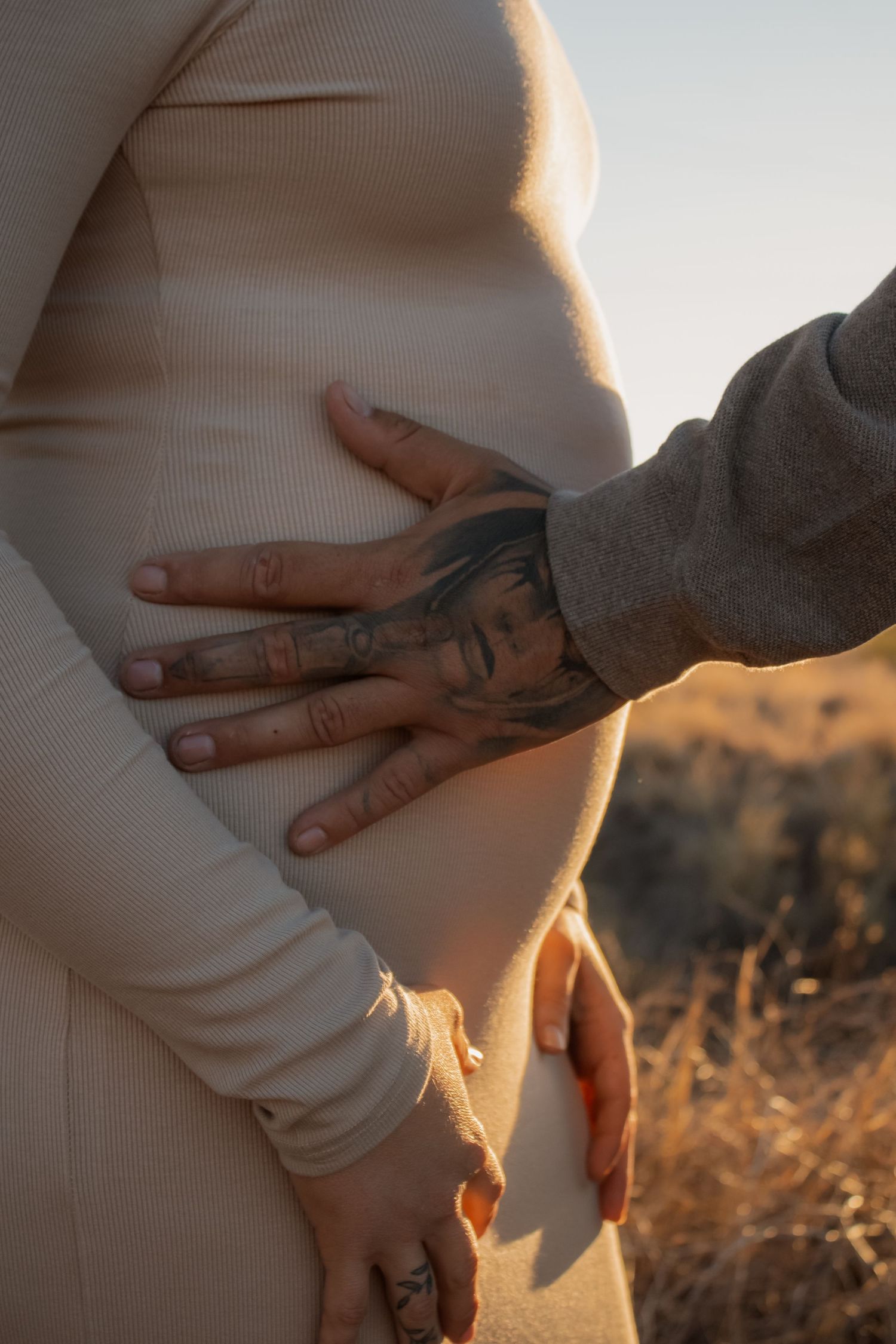 Tattooed hands tenderly cradling a pregnant belly outdoors at golden hour in a beige ribbed dress.