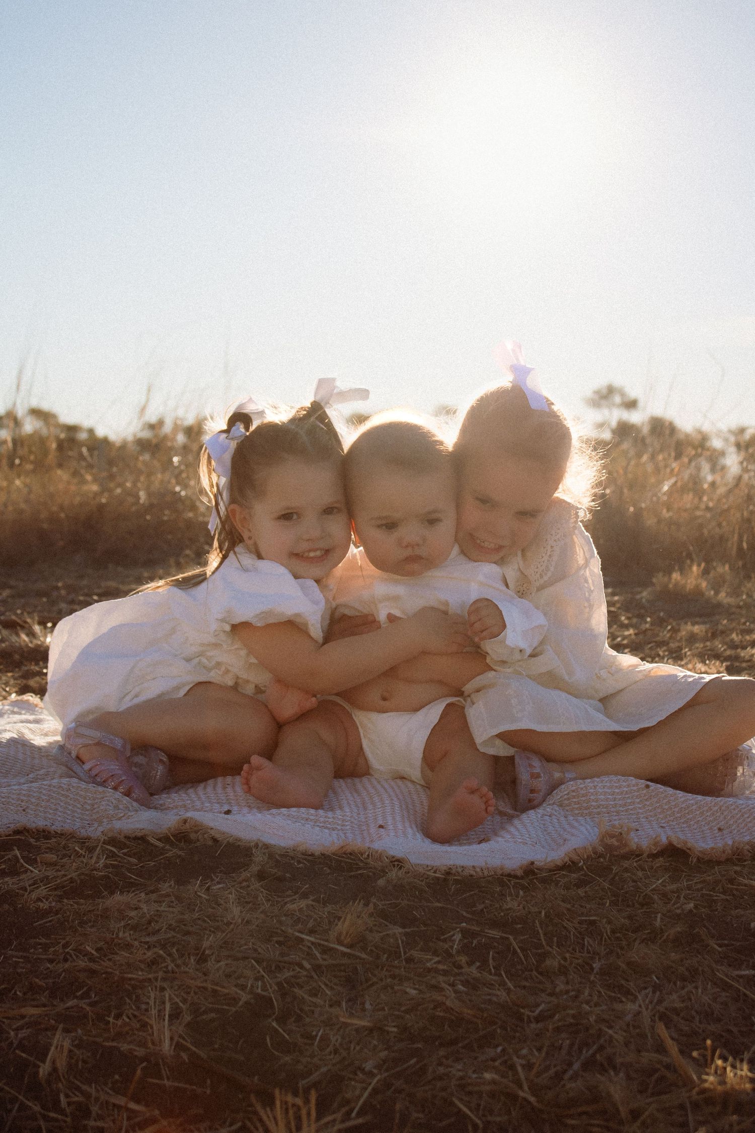 Two sisters  in white dresses sitting close together on a blanket in a sunny field.