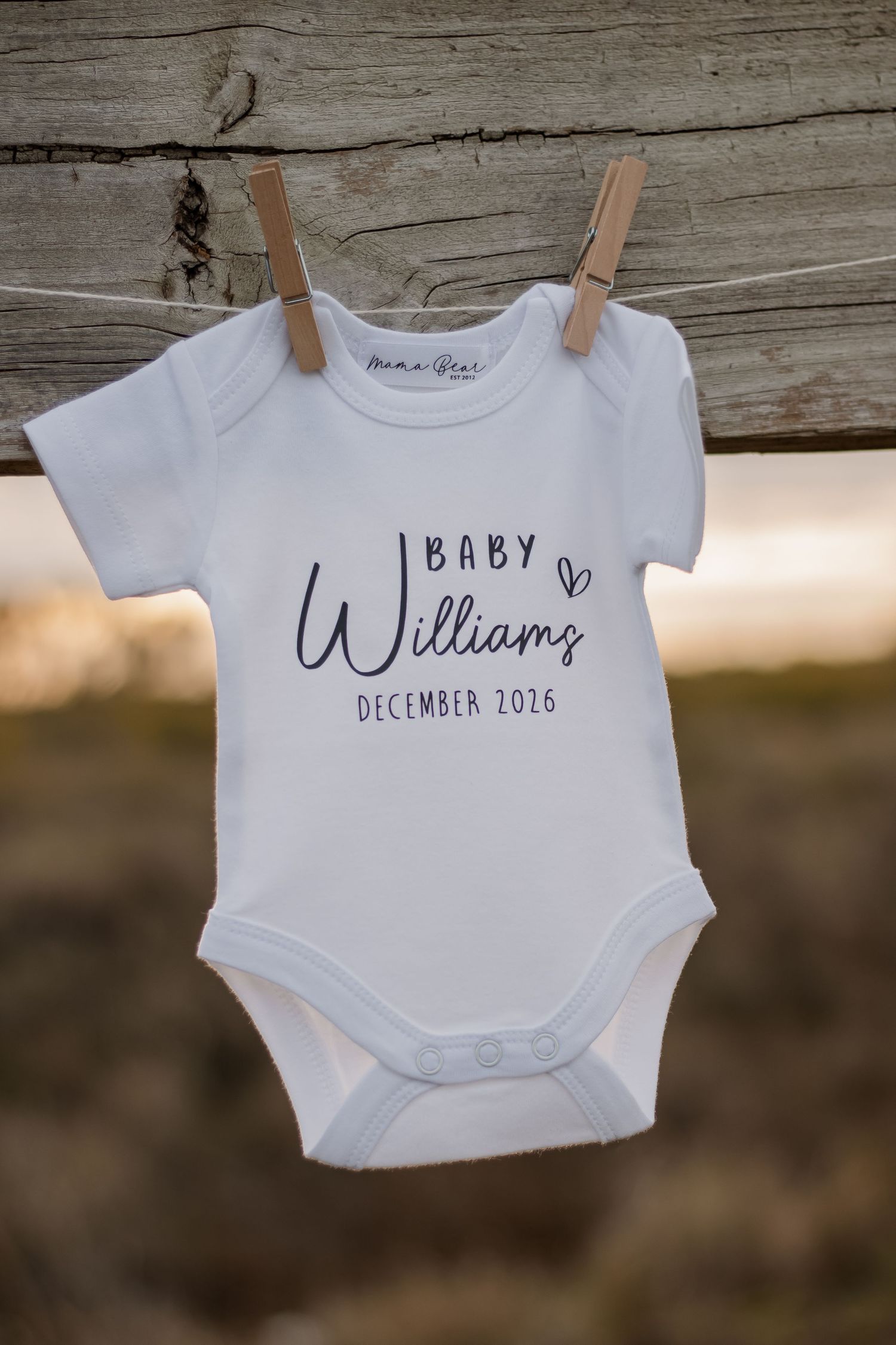 White baby onesie bodysuit hanging on clothesline reading Baby Williams December 2026, pregnancy announcement clothing.