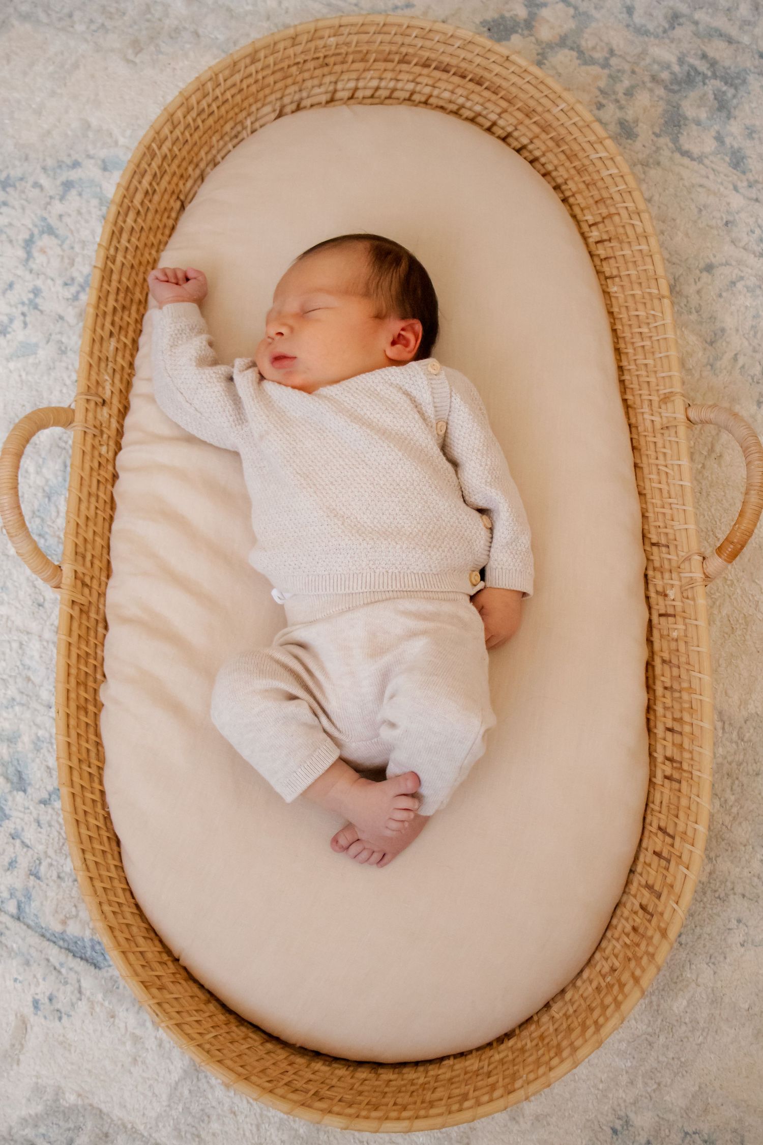 Peaceful newborn baby sleeping in a woven Moses basket on a cream cushion, dressed in a soft white outfit.