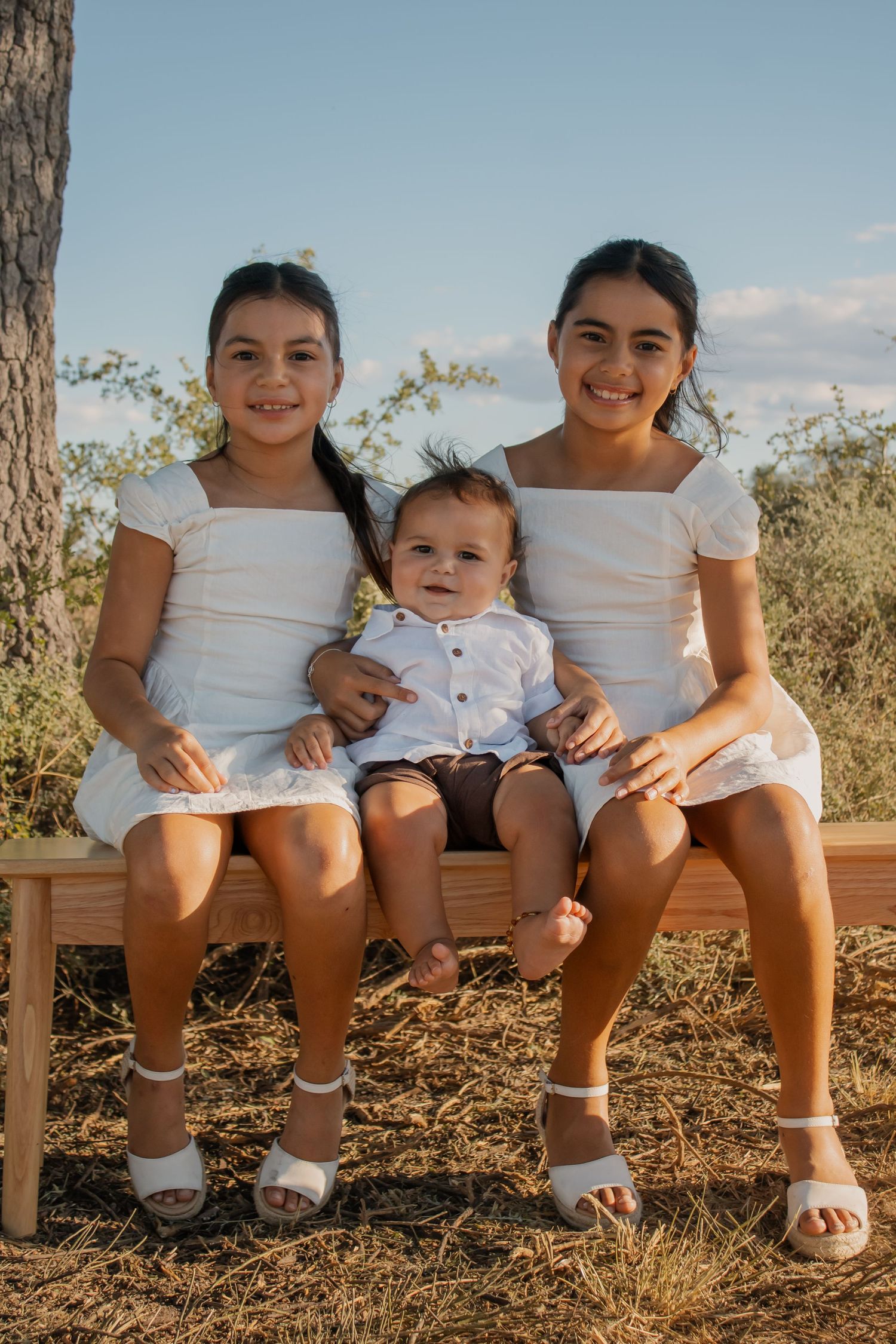 Two girls in white dresses sit on a wooden bench outdoors, holding a baby boy in a white shirt, smiling.