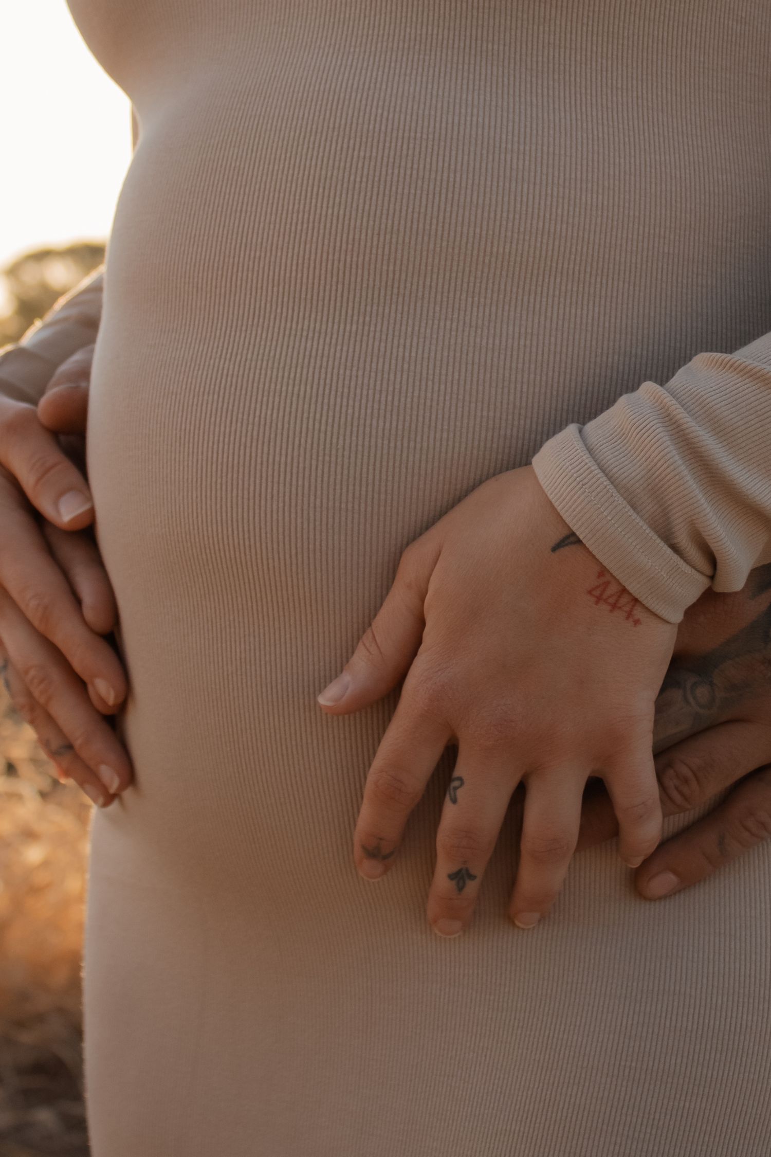 Couple tenderly cradling pregnant belly, tattooed hands visible on beige ribbed maternity dress in warm golden light.