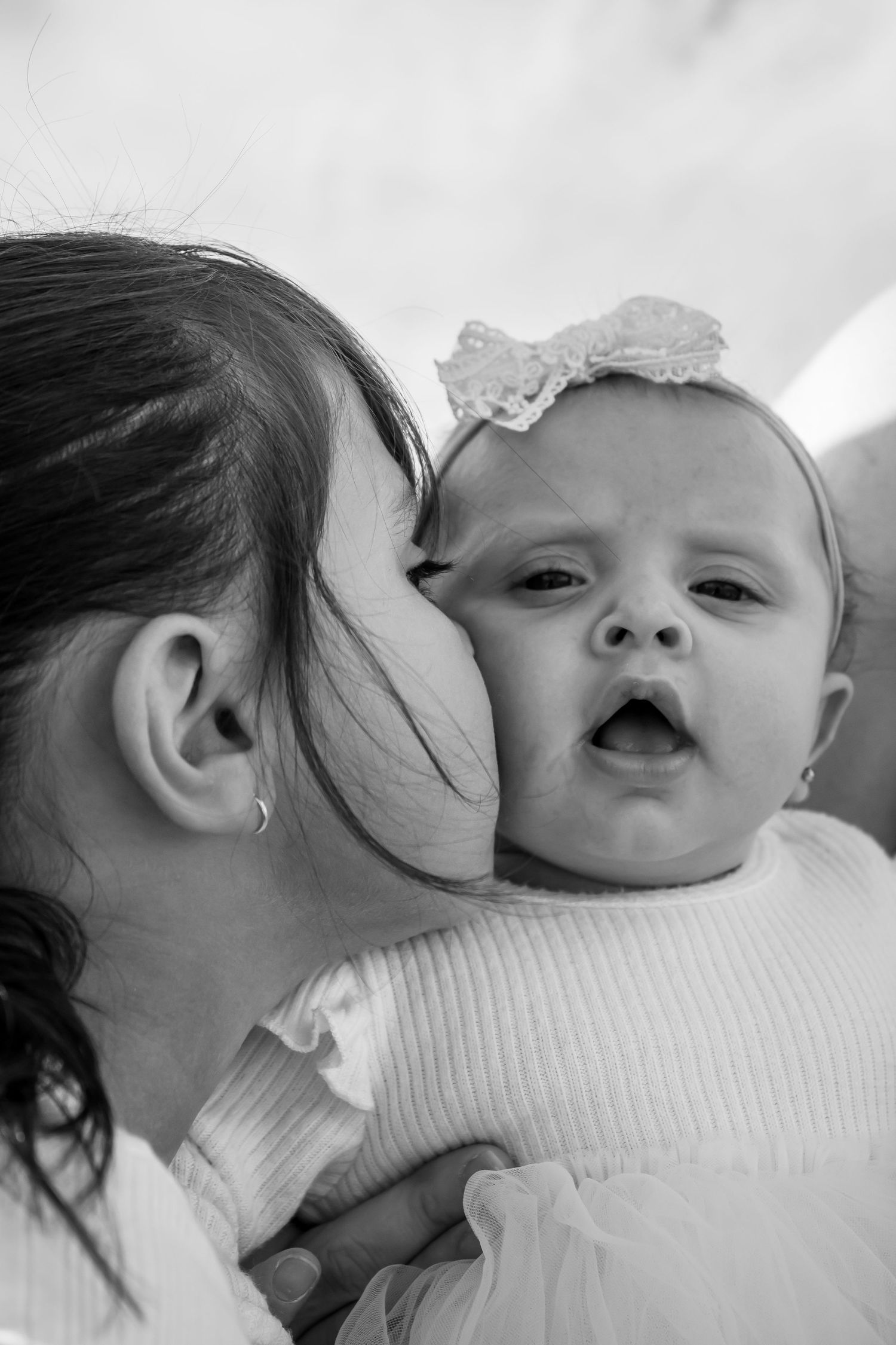 Black and white photo of a young girl kissing a baby wearing a white bow headband and dress.