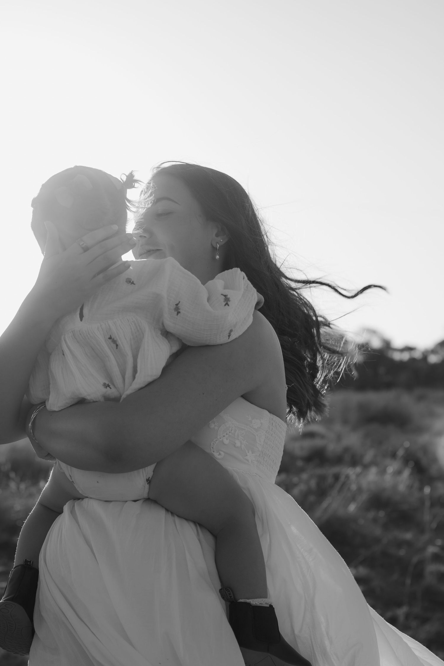 Black and white photo of a joyful woman in a white dress holding and nuzzling a baby outdoors in a field.