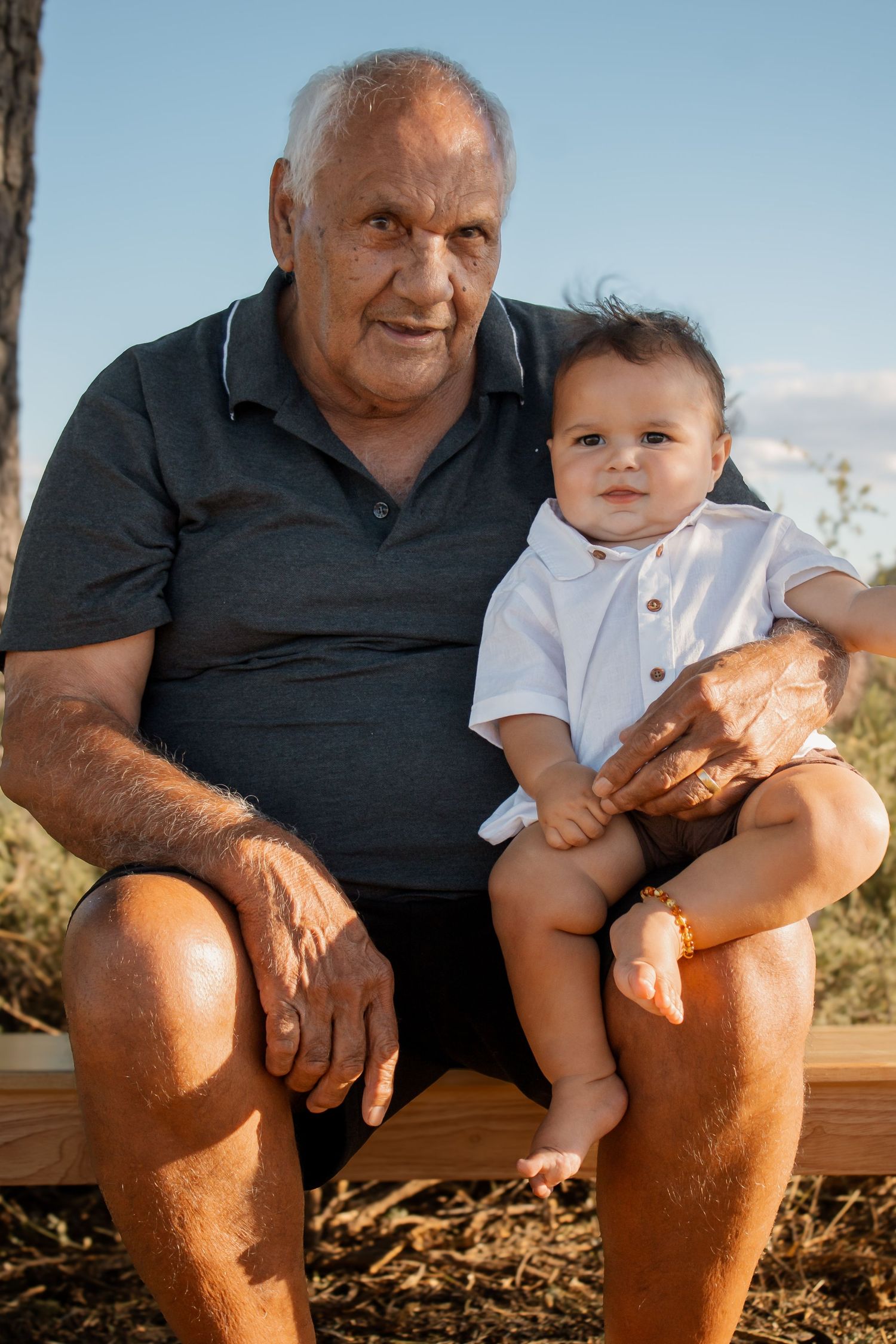 Elderly grandfather sitting outdoors holding a smiling baby on his lap against a blue sky background.