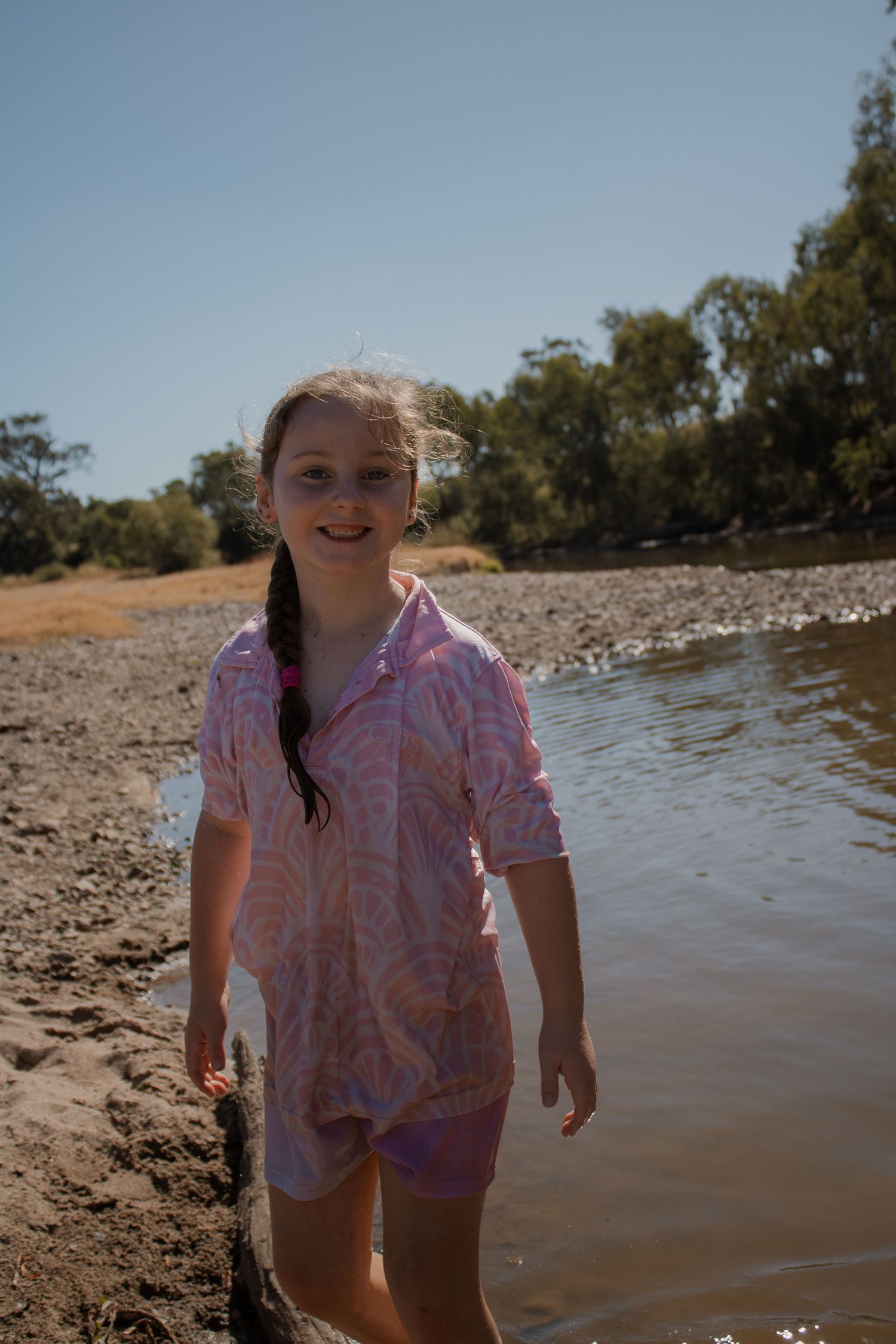 Young girl in pink patterned shirt smiling by a shallow river on a sunny day.
