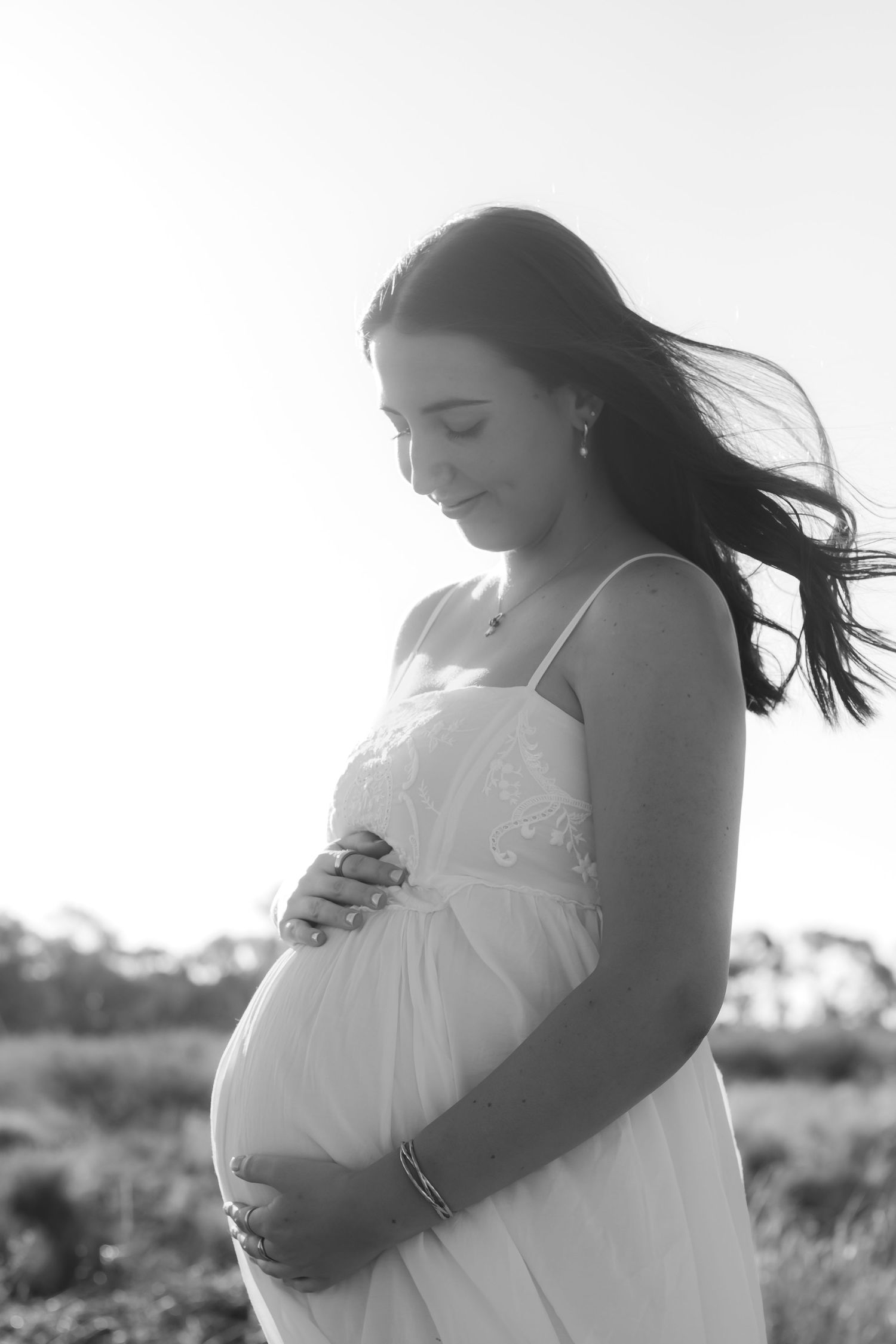 Black and white photo of a pregnant woman in a white dress cradling her baby bump outdoors in a serene field.