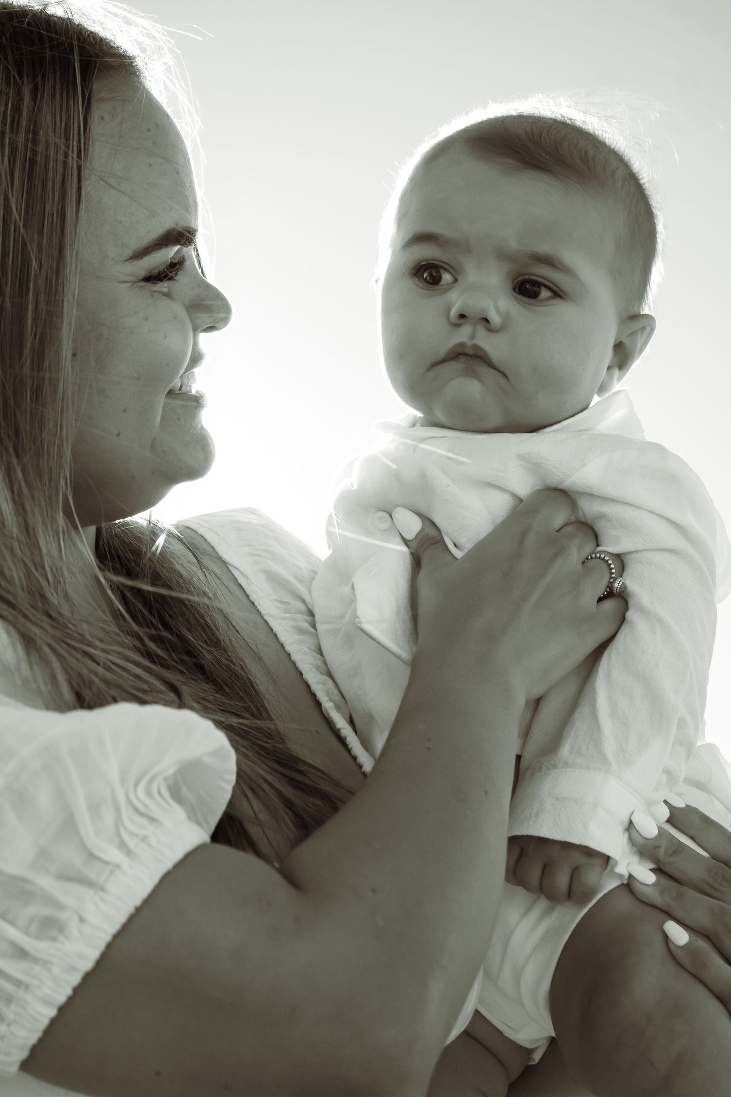 Black and white photo of a smiling mother holding her baby in a white outfit, sharing a tender moment.
