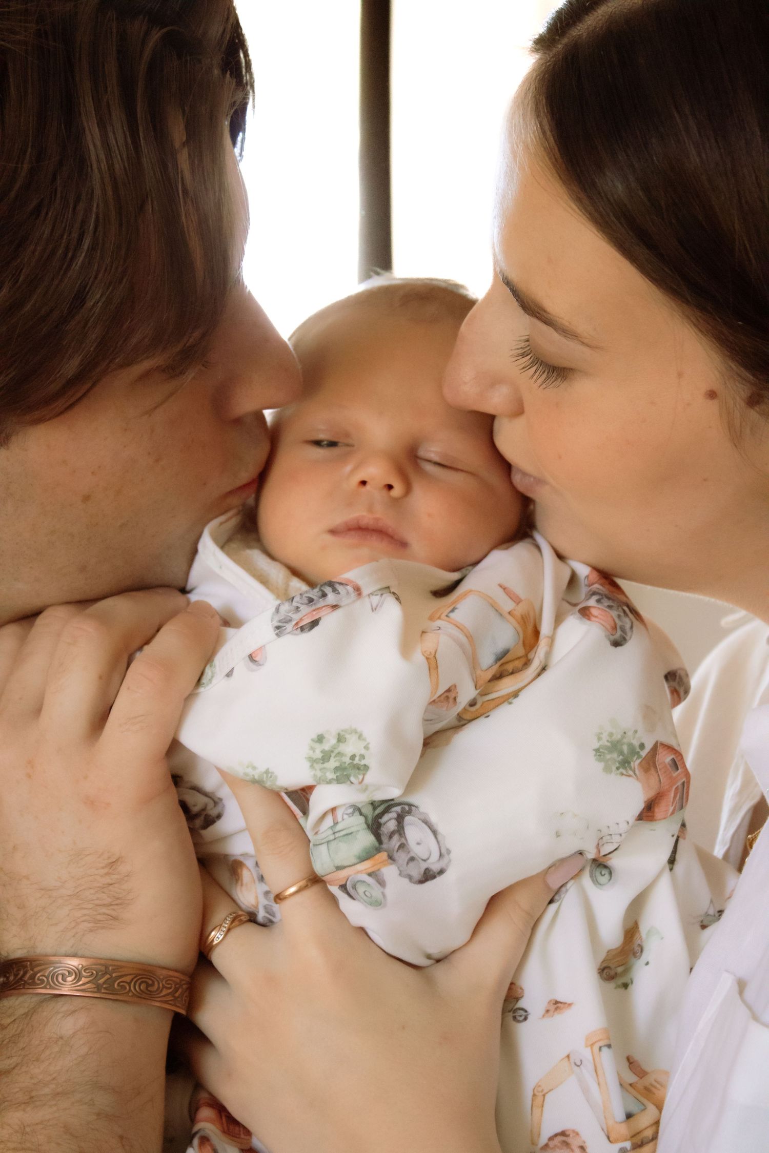 Two parents lovingly kiss their sleeping newborn baby wrapped in a white printed swaddle blanket.