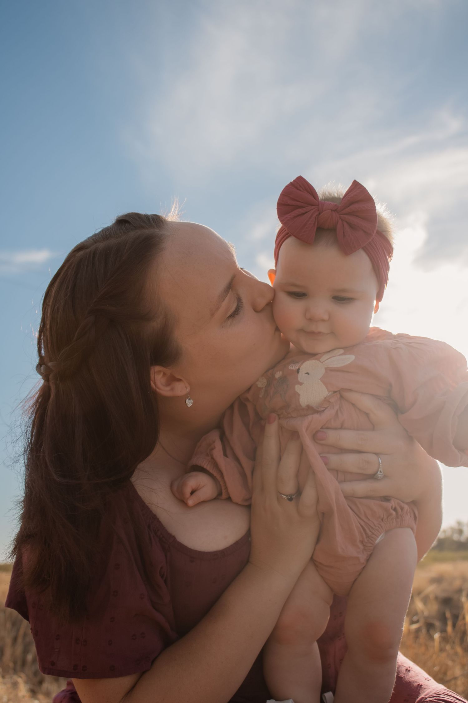 A young mother tenderly kisses her baby girl wearing a pink bow headband outdoors under a sunny blue sky.