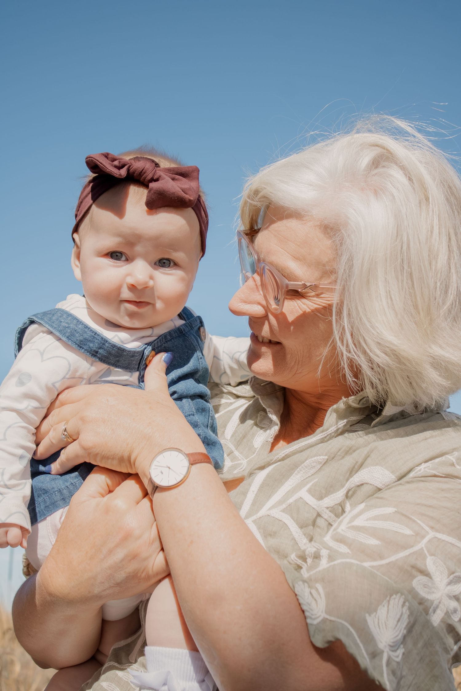 Elderly woman with white hair holding a baby girl wearing a red bow headband outdoors under a blue sky.