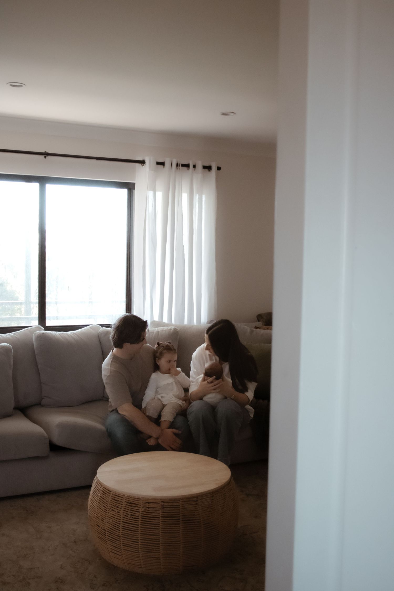 Family of four sitting together on a cozy gray sofa in a bright living room with white curtains.