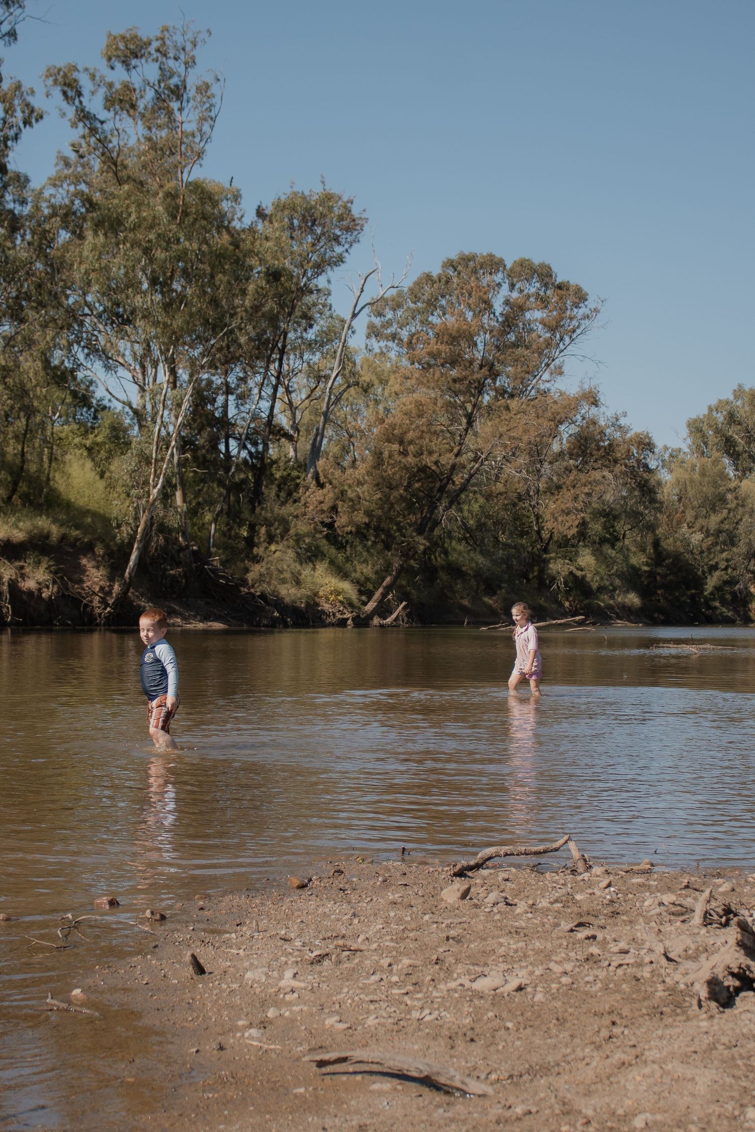 Two children wading in a shallow river surrounded by Australian bush trees on a sunny day.